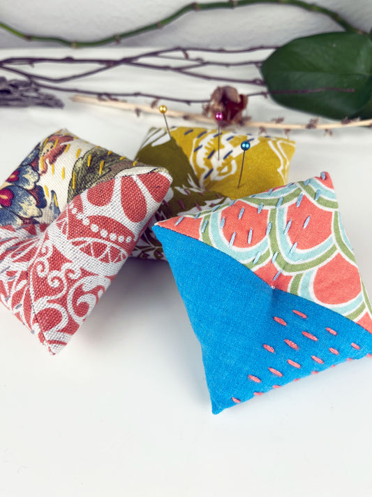 a group of three square pillow shaped pincushions, made from a variety of colorful fabrics with diagonal seams, with sashiko stitching in contrasting colors, on a white counter with dried twigs seedpods and a pothos plant in the background
