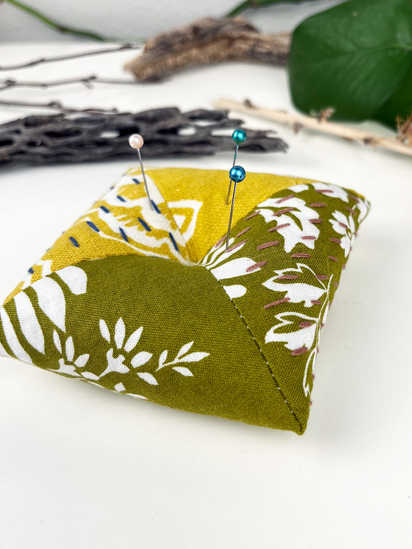 a square pillow shaped pincushion made from olive and mustard floral fabric, with sashiko stitching in contrasting colors, on a white counter with dried twigs seedpods and a pothos plant in the background