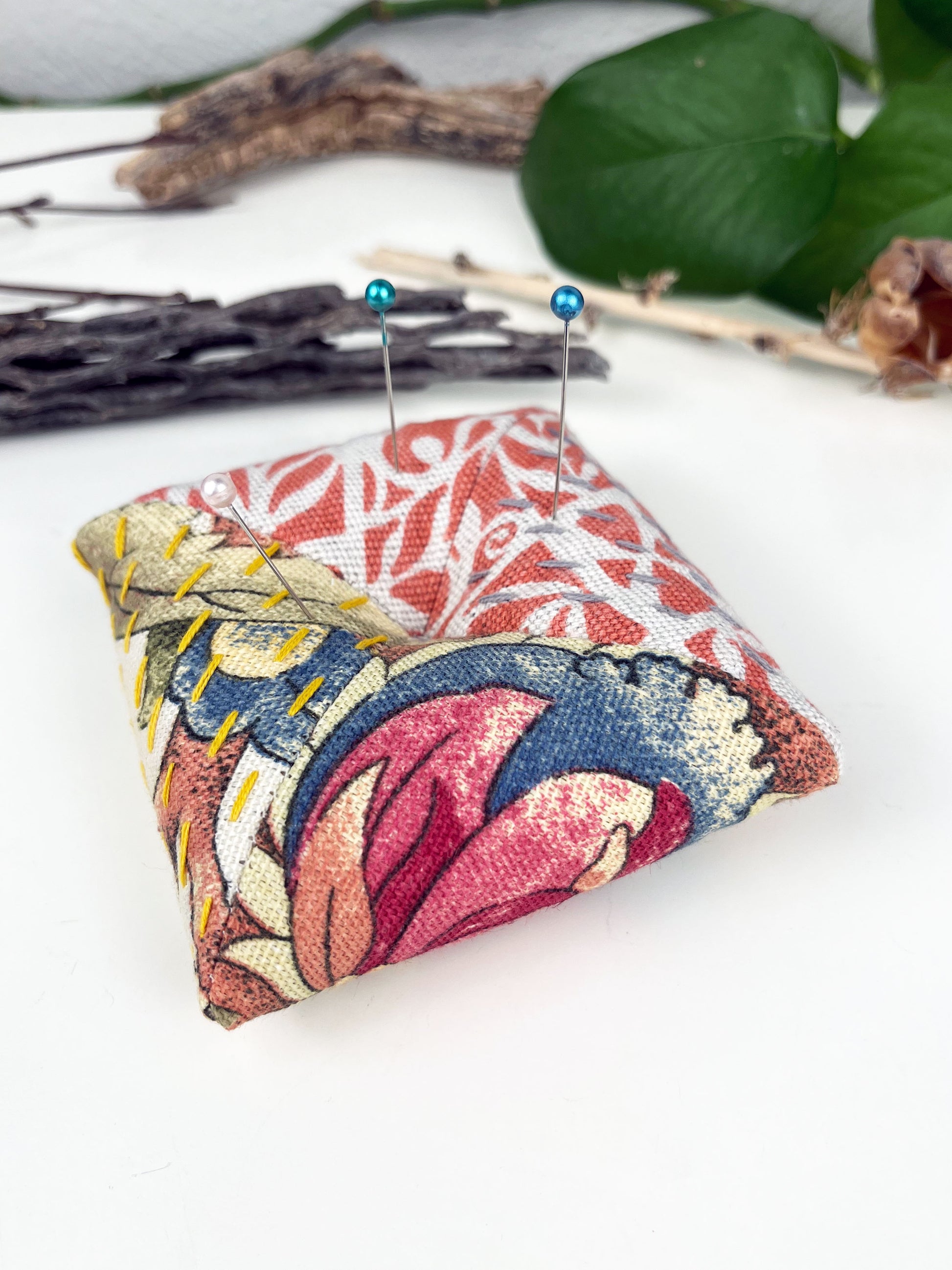 a square pillow shaped pincushion made from repurposed orange and floral upholstery fabric, with sashiko stitching in contrasting colors, on a white counter with dried twigs seedpods and a pothos plant in the background