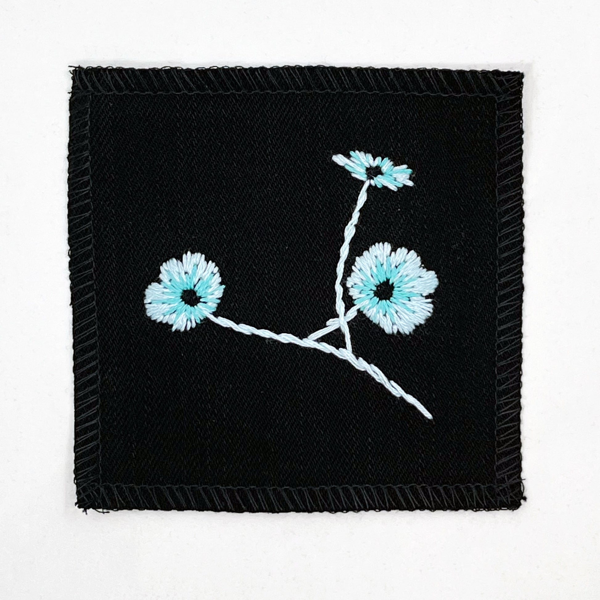 a black colored square patch, hand embroidered with three poppies on a stem, in shades of blue, with overlocked edges, on a white background