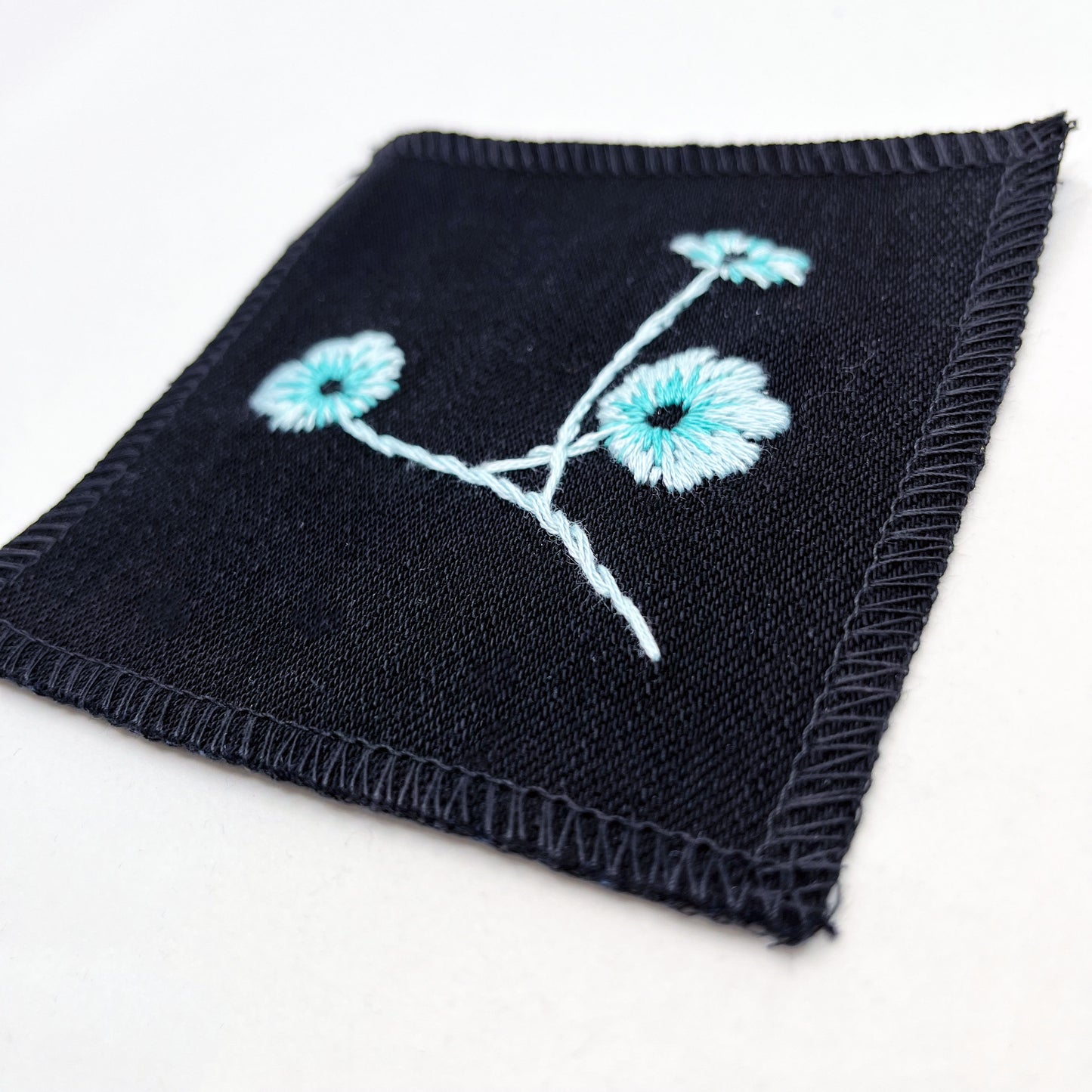 a close up angled view of a black colored square patch, hand embroidered with three poppies on a stem, in shades of blue, with overlocked edges, on a white background