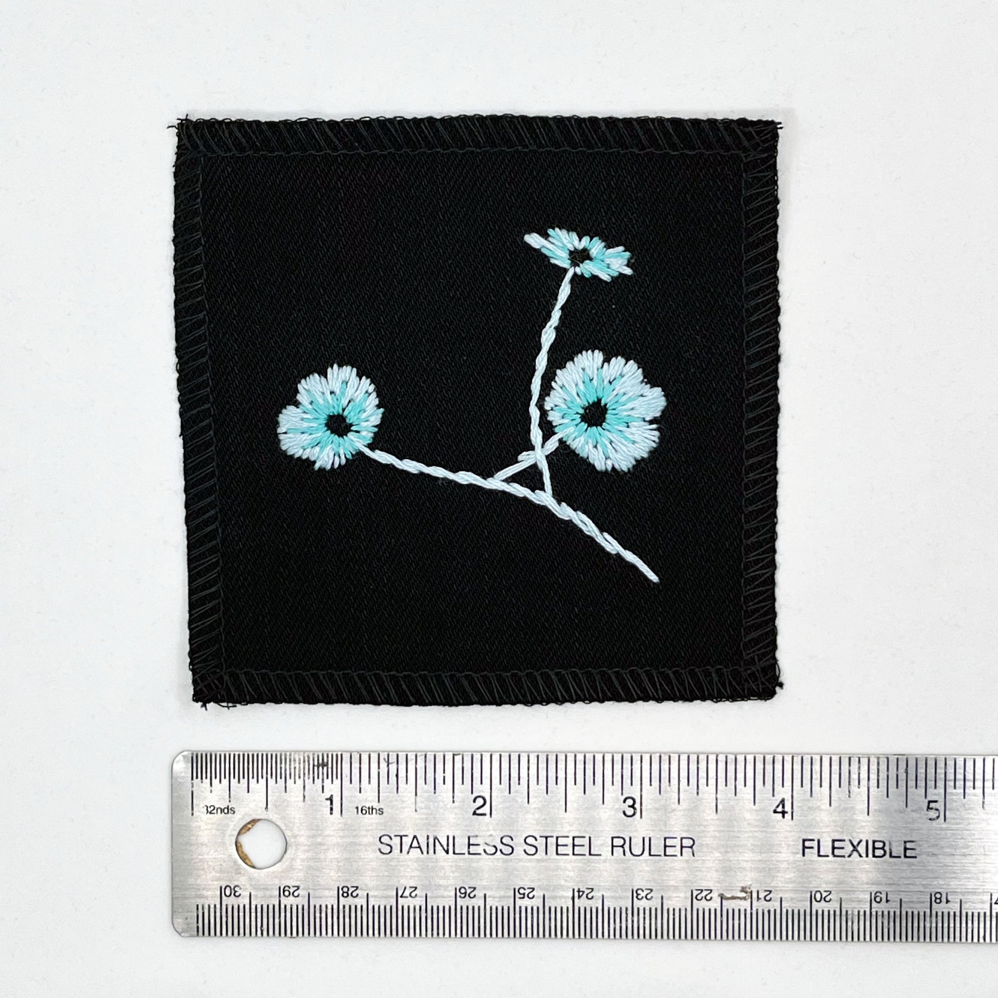 a black colored square patch, hand embroidered with three poppies on a stem, in shades of blue, with overlocked edges, placed next to a metal ruler to show a width of four inches, on a white background