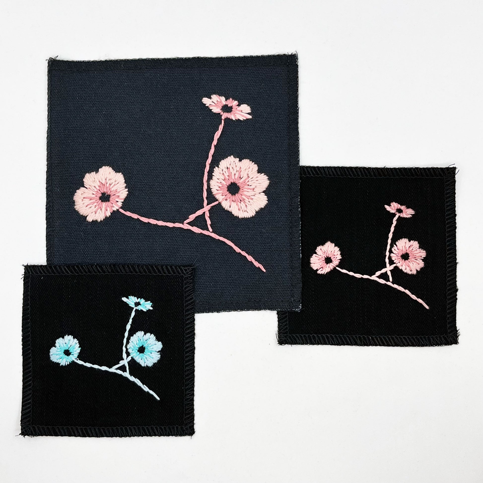 a group of square patches in black fabric, hand embroidered with three poppies on stems, some in shades of pink, some in shades of blue