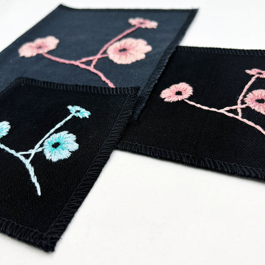 a group of square patches in black fabric, hand embroidered with three poppies on stems, some in shades of pink, some in shades of blue