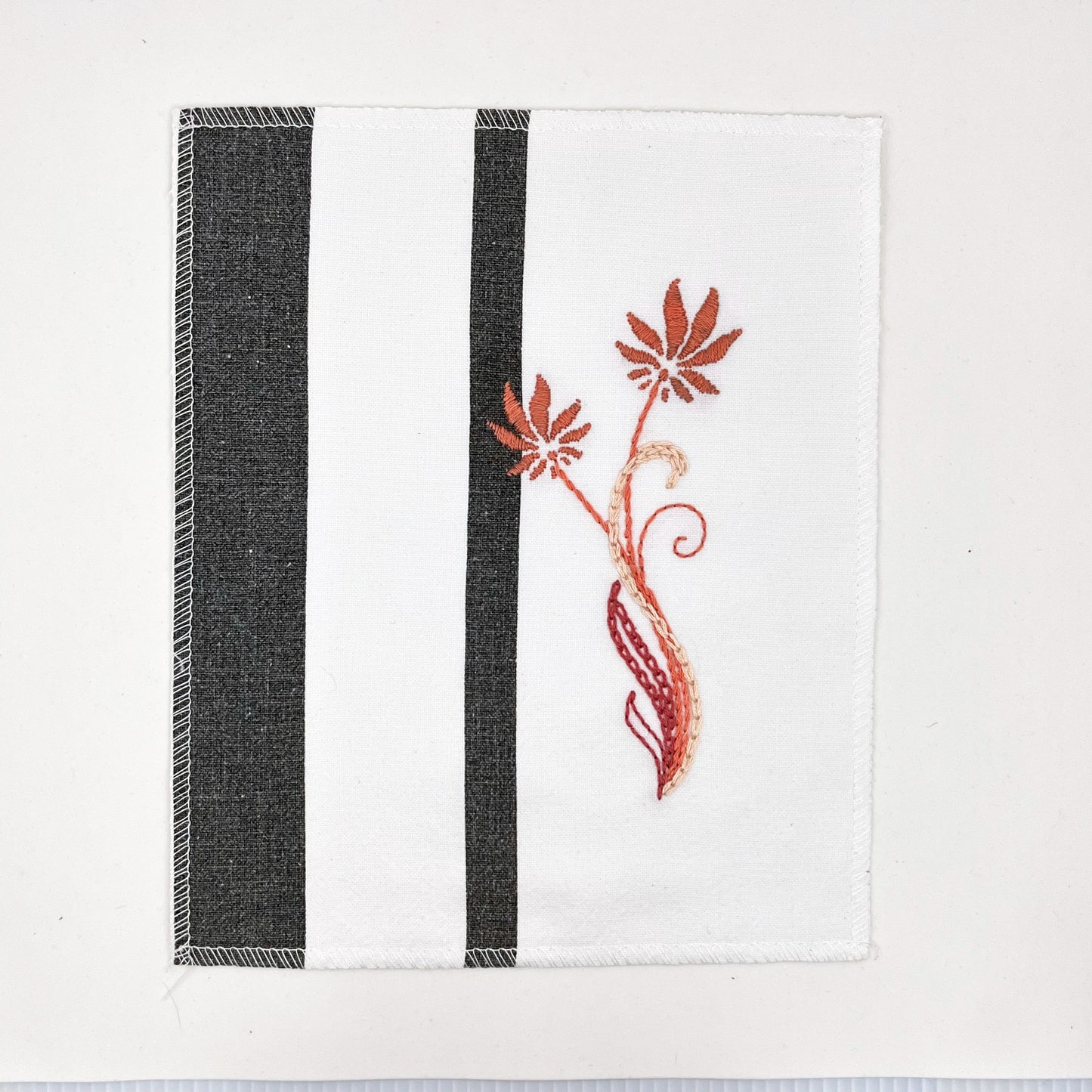 a fabric wall hanging made out of white fabric with black vertical stripes, hand stitched with flowers in shades of coral with long curvy stems in chainstitch, backstitch and stem stitch, on a white background