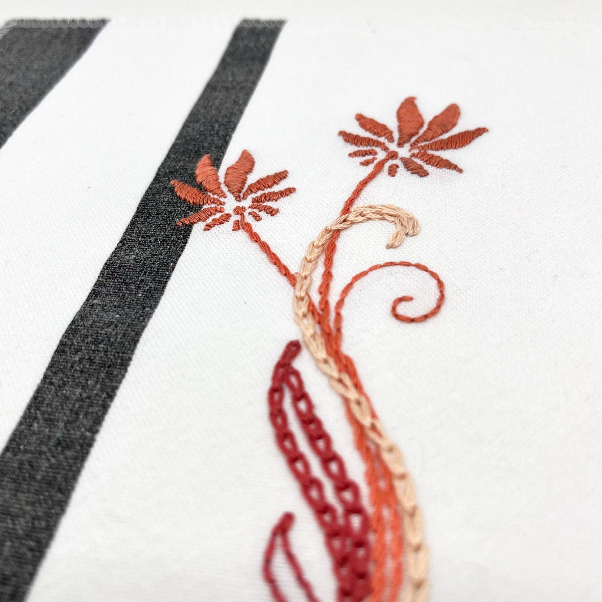 close up angled view of a fabric wall hanging made out of white fabric with black vertical stripes, hand stitched with flowers in shades of coral with long curvy stems in chainstitch, backstitch and stem stitch, on a white background