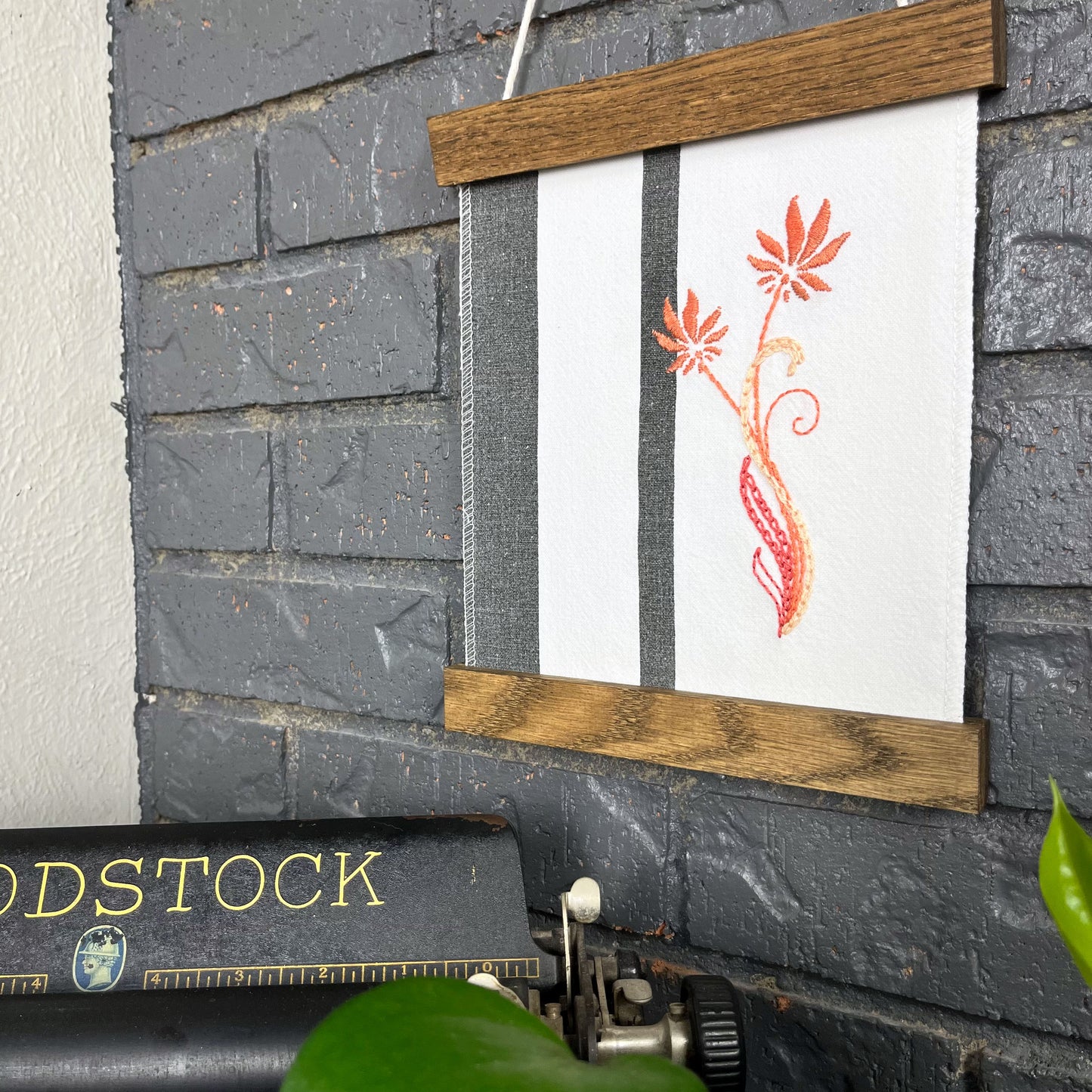 a fabric wall hanging in a wood magnetic frame on a grey wall, made out of white fabric with black vertical stripes, hand stitched with coral flowers with long curvy stems, a typewriter and pothos plant peek out at the bottom of the frame