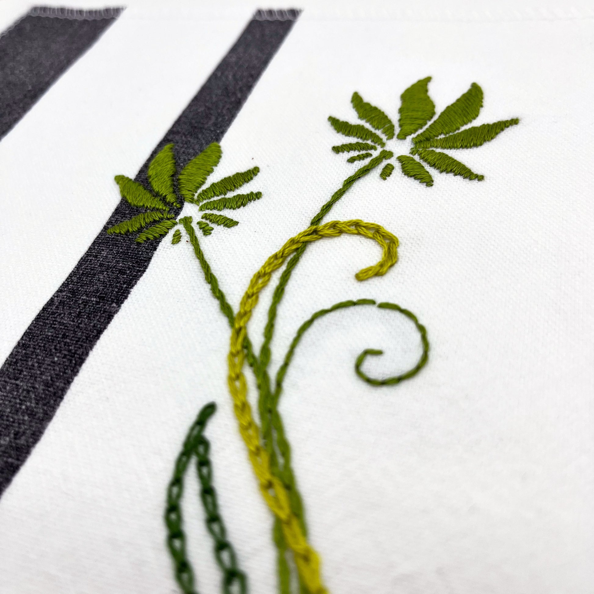 close up angled view of a fabric wall hanging made out of white fabric with black vertical stripes, hand stitched with flowers in shades of green with long curvy stems in chainstitch, backstitch and stem stitch, on a white background