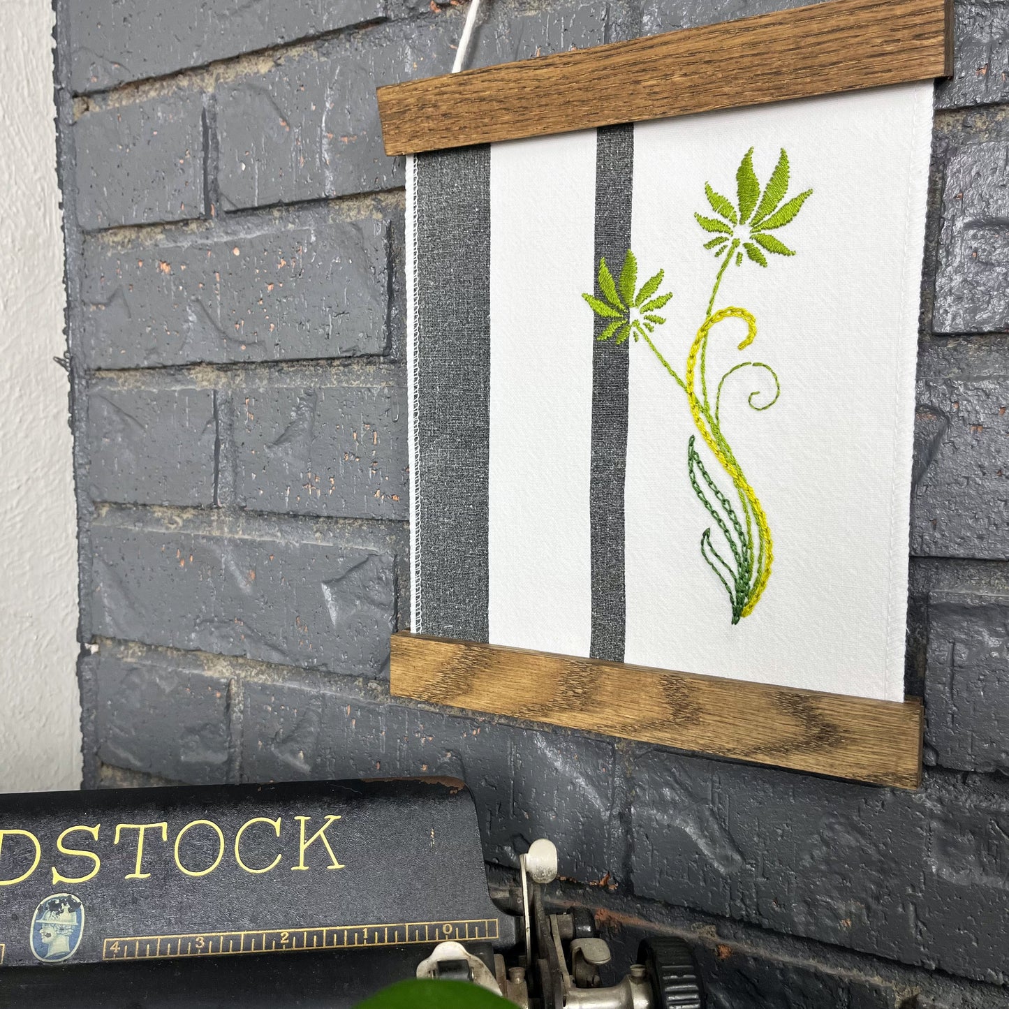 a fabric wall hanging in a wood magnetic frame on a grey wall, made out of white fabric with black vertical stripes, hand stitched with green flowers with long curvy stems, a typewriter and pothos plant peek out at the bottom of the frame