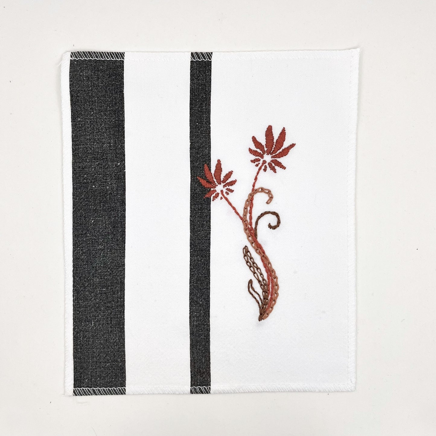 a fabric wall hanging made out of white fabric with black vertical stripes, hand stitched with flowers in shades of mauve with long curvy stems in chainstitch, backstitch and stem stitch, on a white background