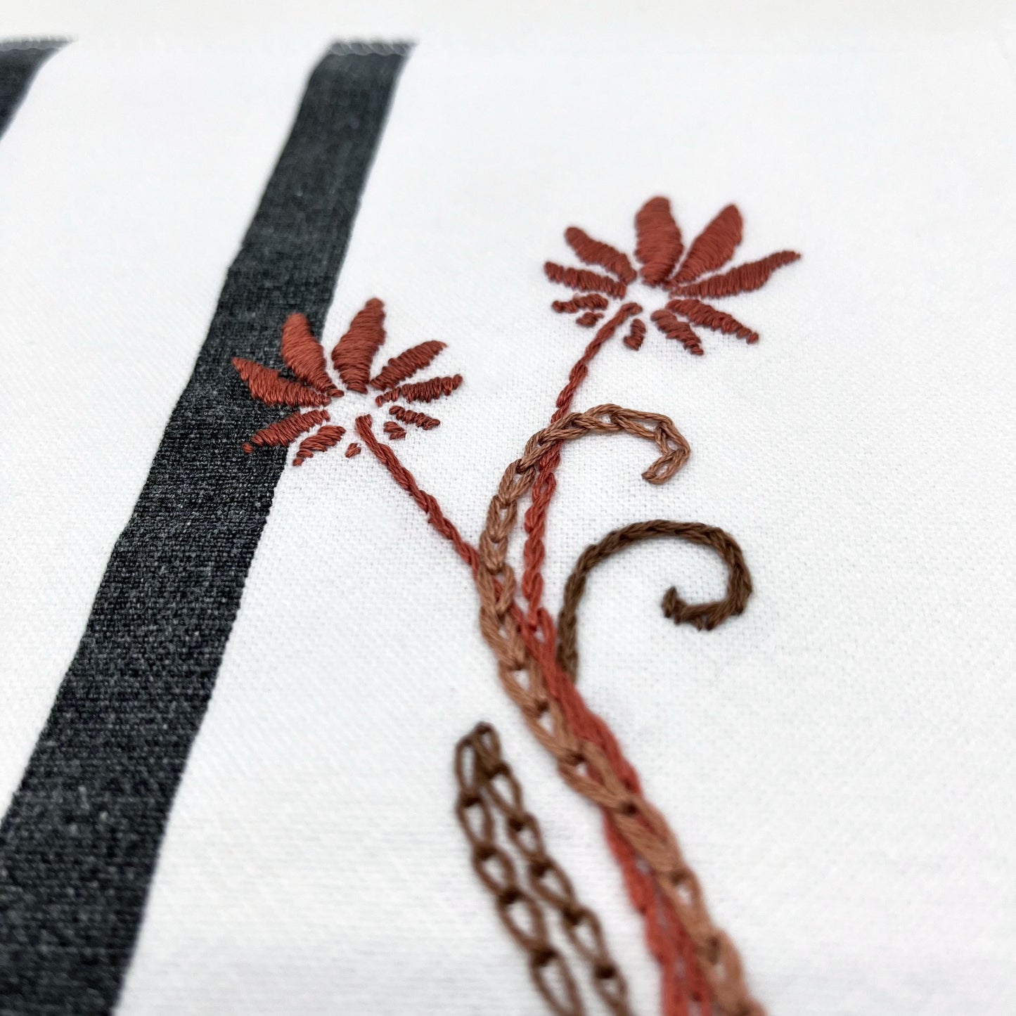 close up angled view of a fabric wall hanging made out of white fabric with black vertical stripes, hand stitched with flowers in shades of mauve with long curvy stems in chainstitch, backstitch and stem stitch, on a white background
