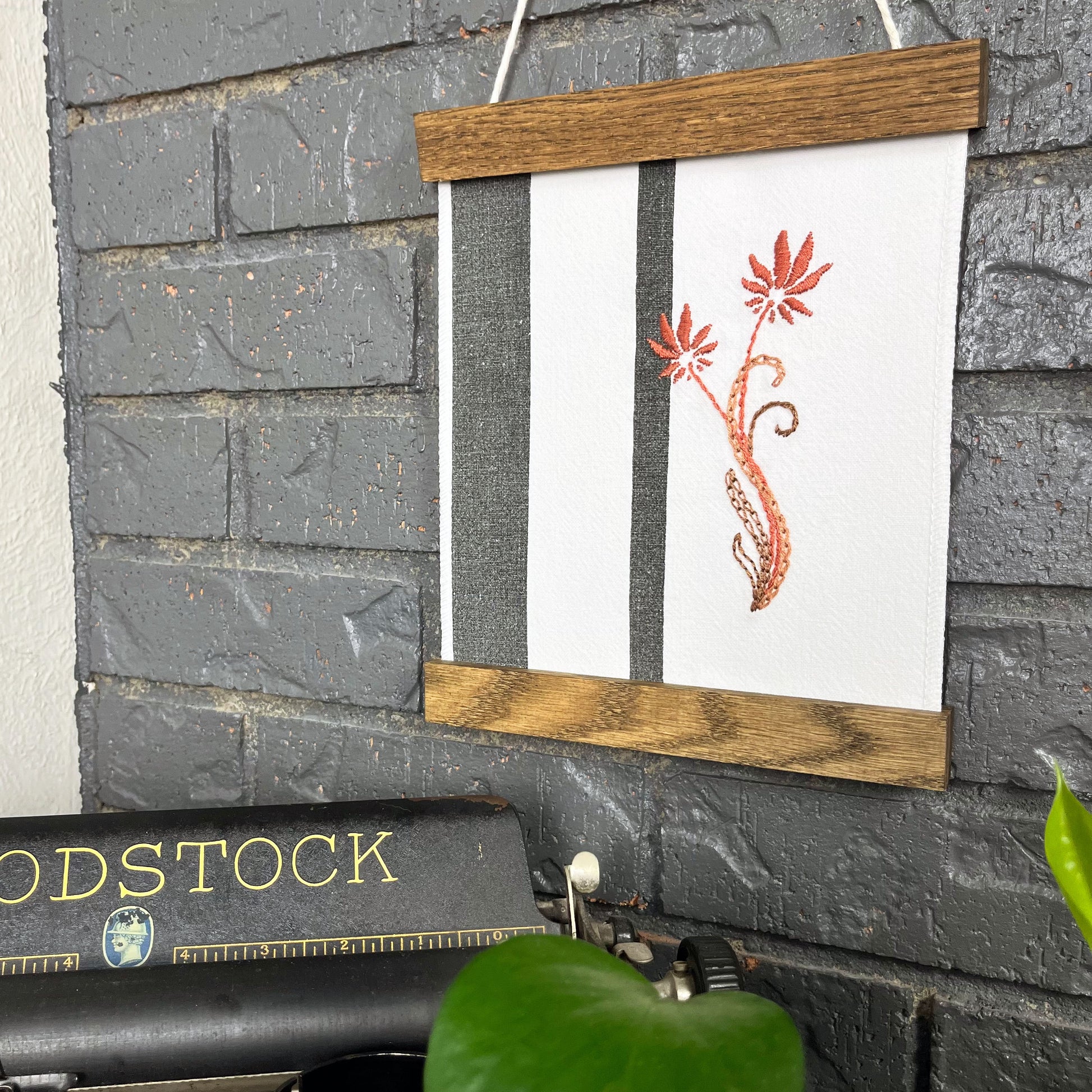 a fabric wall hanging in a wood magnetic frame on a grey wall, made out of white fabric with black vertical stripes, hand stitched with mauve flowers with long curvy stems, a typewriter and pothos plant peek out at the bottom of the frame