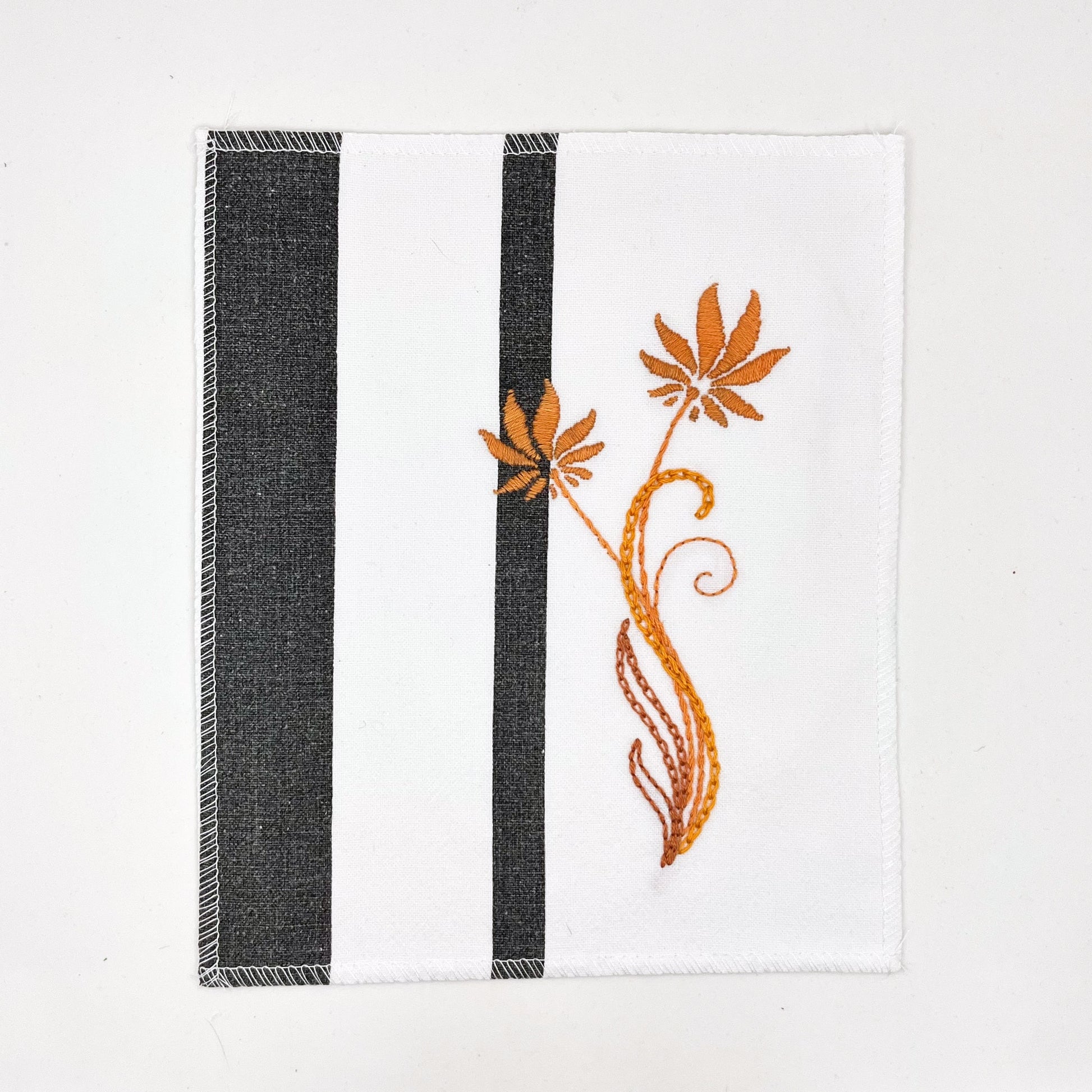 a fabric wall hanging made out of white fabric with black vertical stripes, hand stitched with orange flowers with long curvy stems in chainstitch, backstitch and stem stitch, on a white background