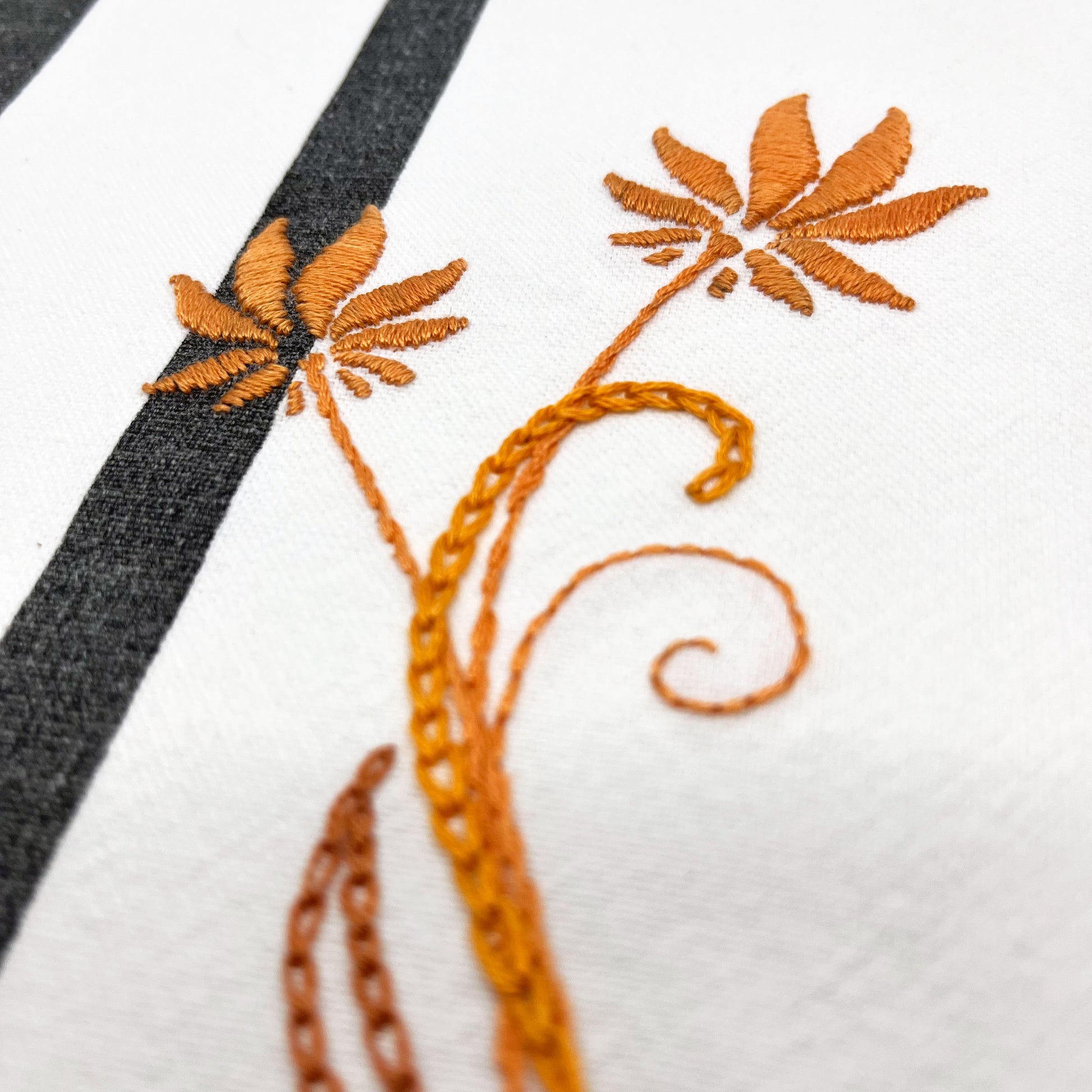 close up angled view of a fabric wall hanging made out of white fabric with black vertical stripes, hand stitched with flowers in shades of orange with long curvy stems in chainstitch, backstitch and stem stitch, on a white background