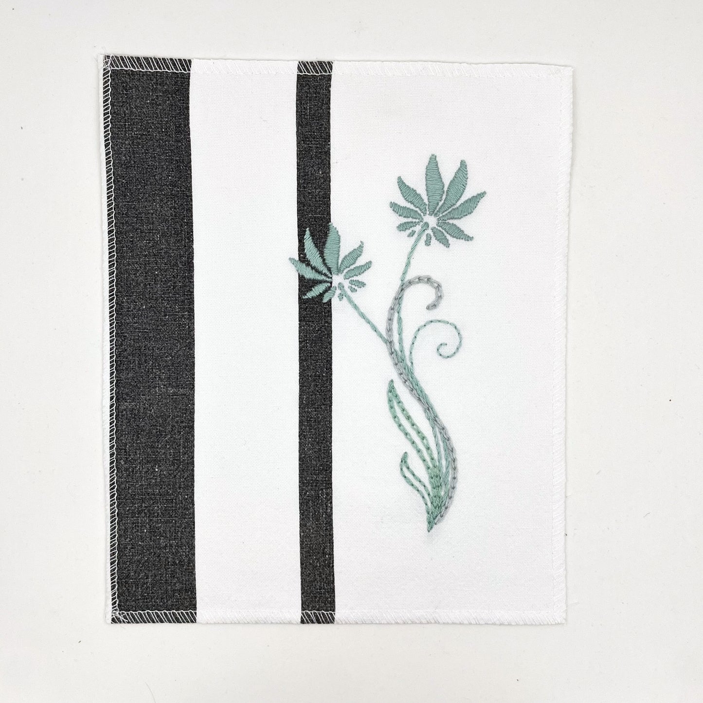 a fabric wall hanging made out of white fabric with black vertical stripes, hand stitched with flowers in shades of sage blue with long curvy stems in chainstitch, backstitch and stem stitch, on a white background