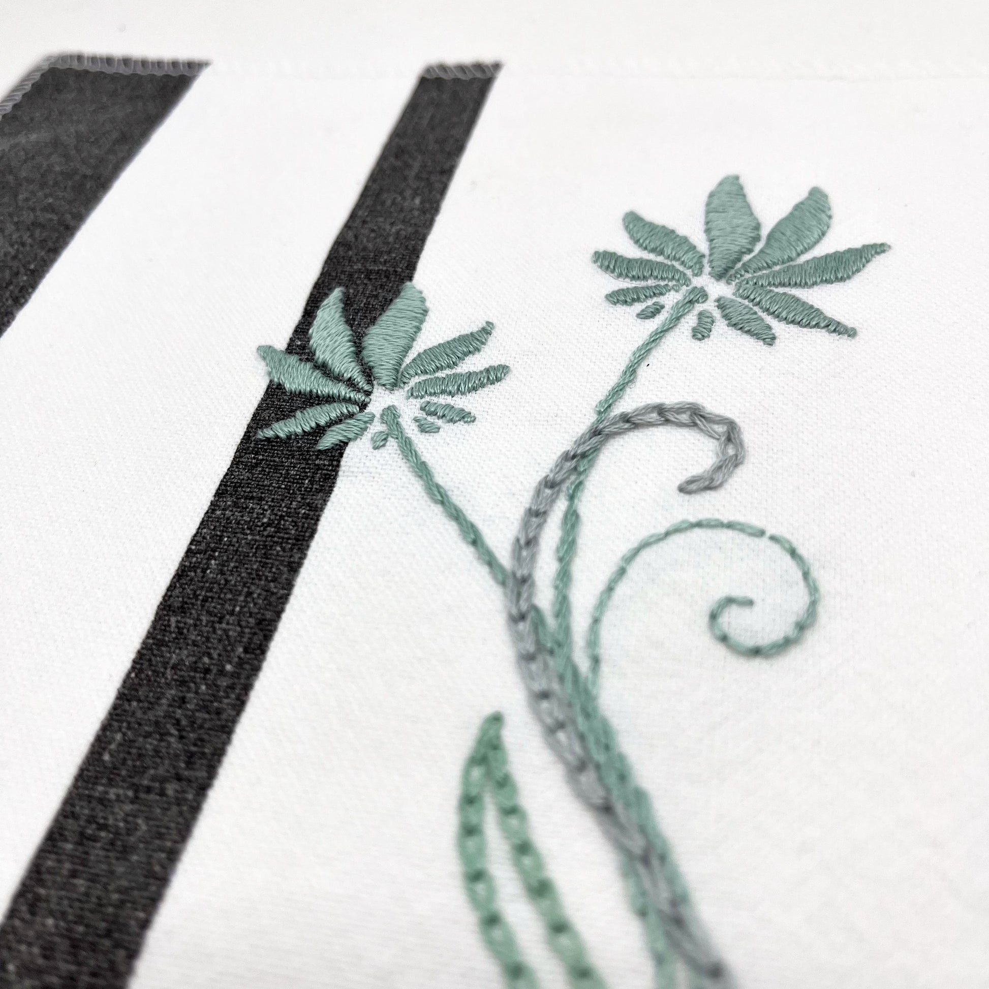 close up angled view of a fabric wall hanging made out of white fabric with black vertical stripes, hand stitched with flowers in shades of sage with long curvy stems in chainstitch, backstitch and stem stitch, on a white background
