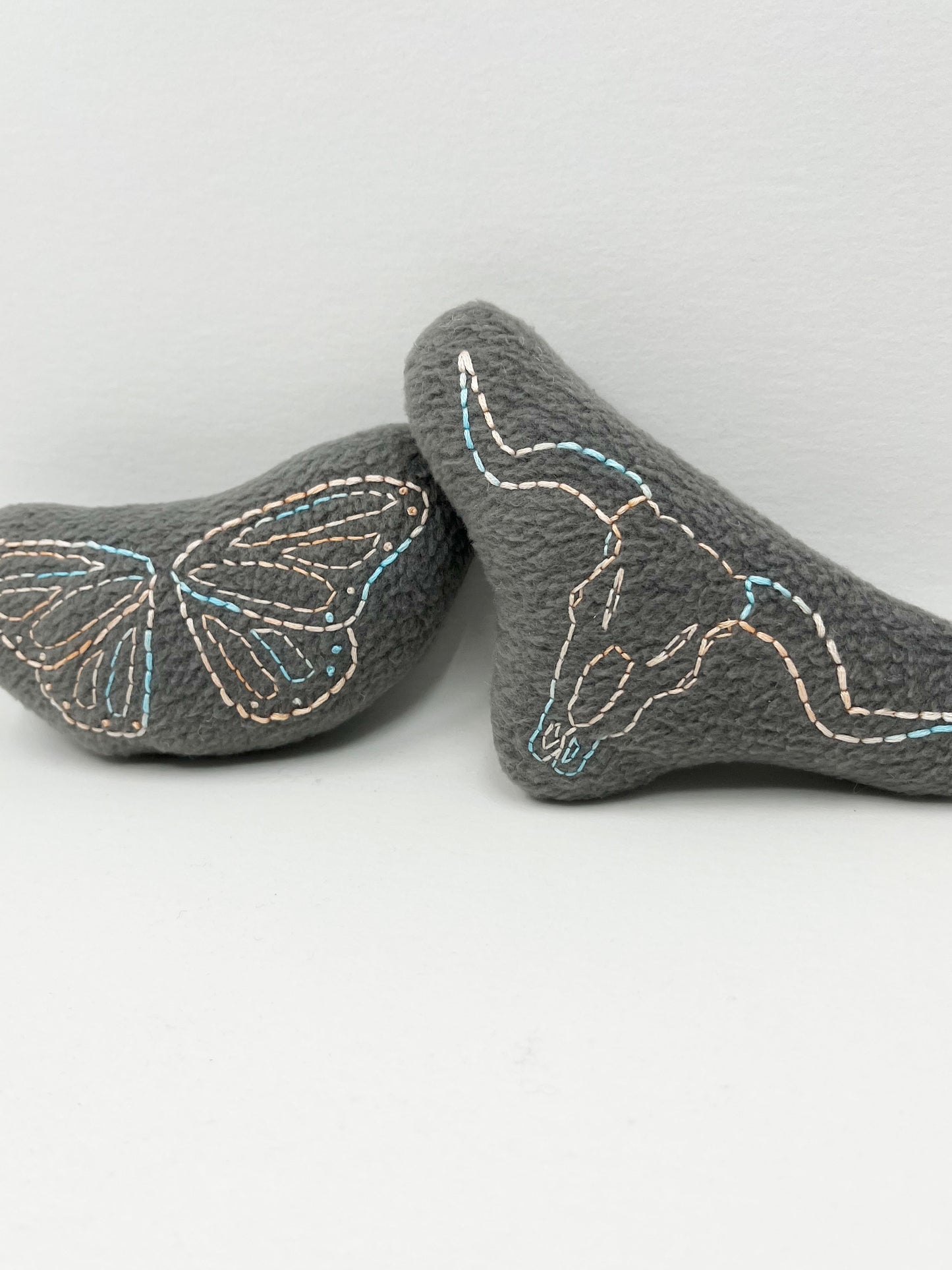 two grey stuffies hand embroidered in peach and sky blue variegated thread with a longhorn skull and butterfly wings on a white background