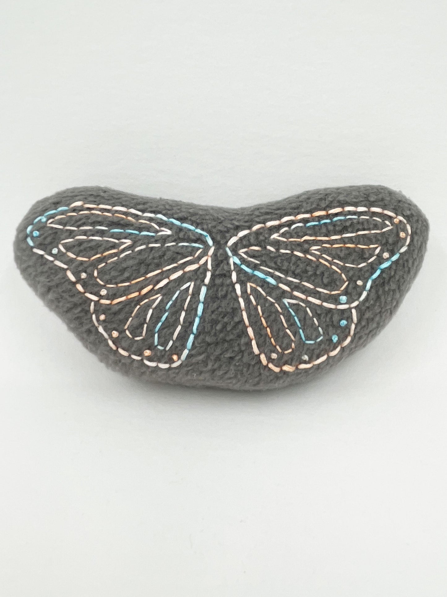 a grey fleece stuffed animal with butterfly wings hand embroidered on it in peach and sky blue variegated thread with backstitch and french knots on a white background