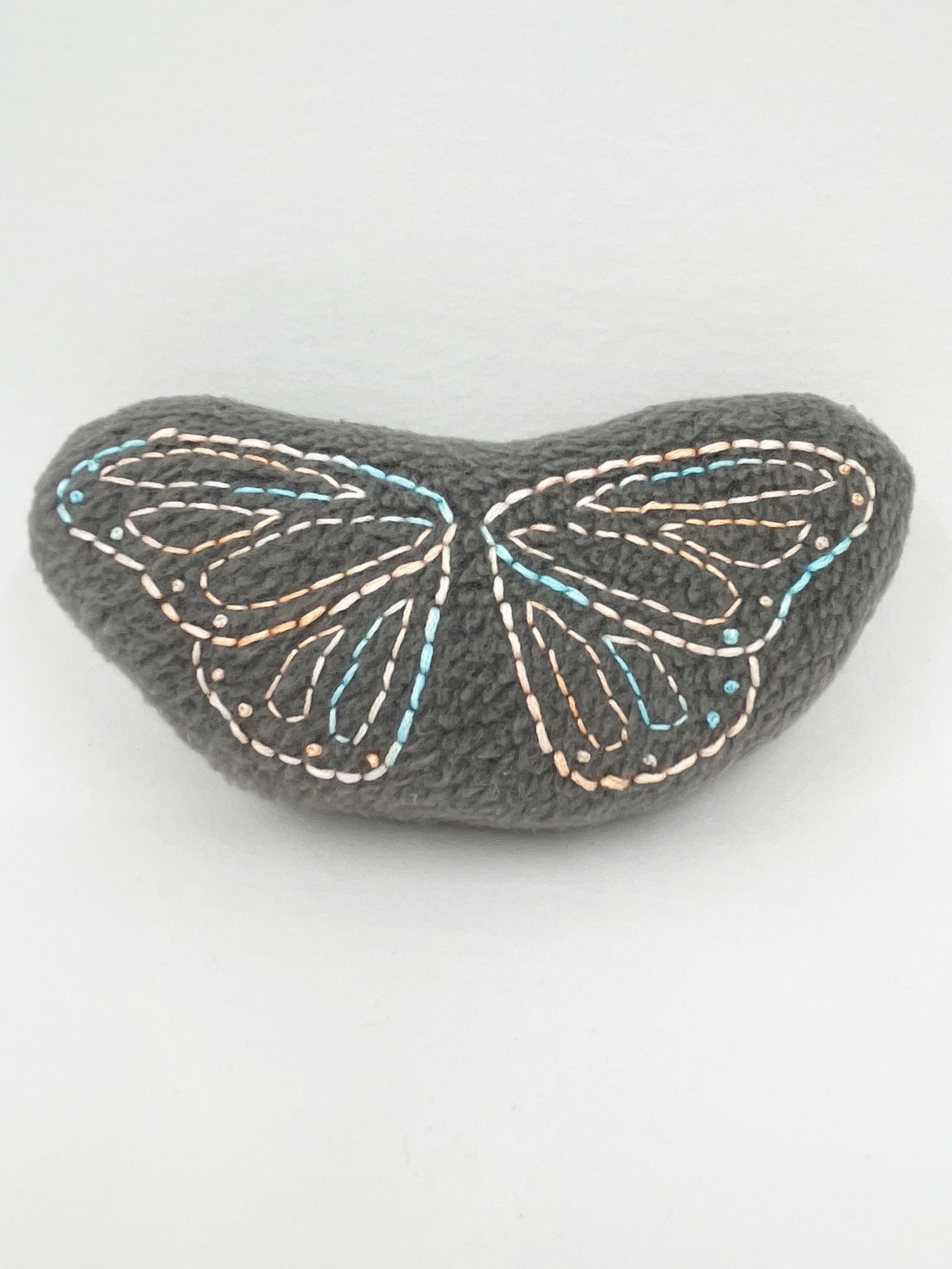 a grey fleece stuffed animal with butterfly wings hand embroidered on it in peach and sky blue variegated thread with backstitch and french knots on a white background