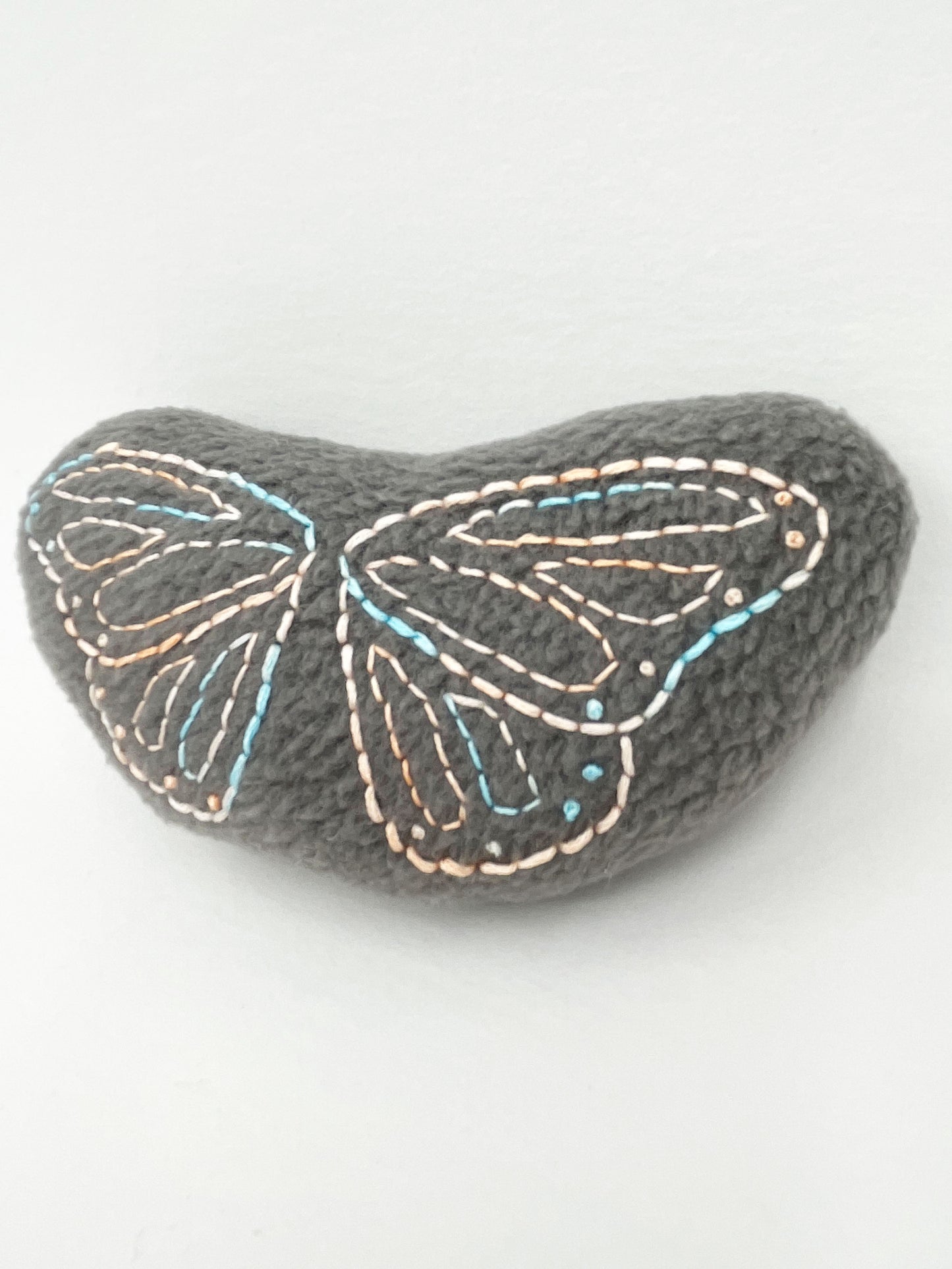 a grey fleece stuffed animal with butterfly wings hand embroidered on it in peach and sky blue variegated thread with backstitch and french knots on a white background