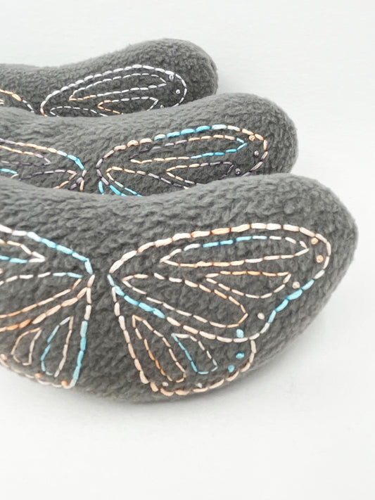 three grey fleece stuffed animals lined up from front to back, with butterfly wings hand embroidered on them in peach and sky blue variegated thread with backstitch and french knots