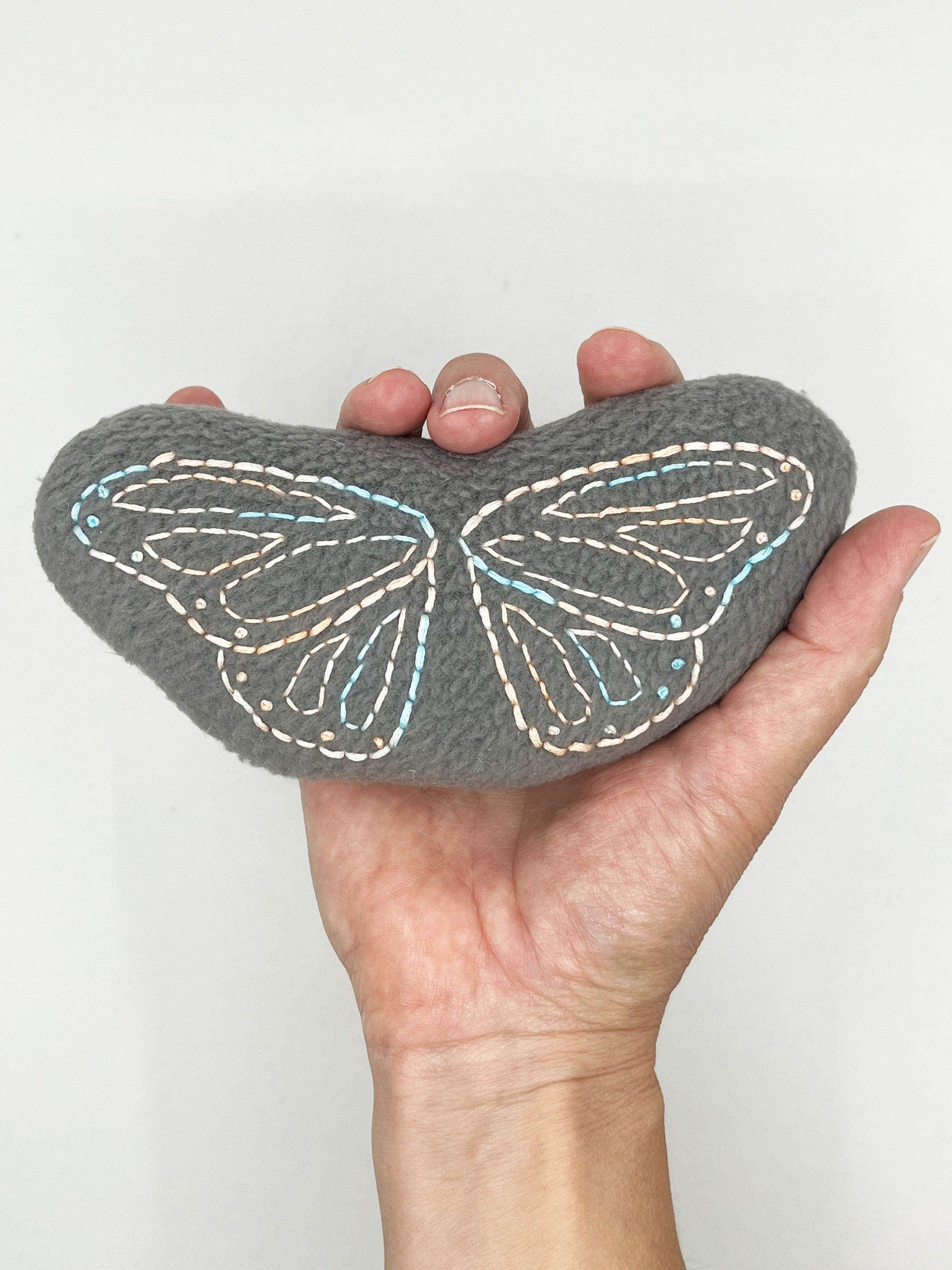 a hand holding up a grey fleece stuffed animal with butterfly wings hand embroidered on it in peach and sky blue variegated thread with backstitch and french knots