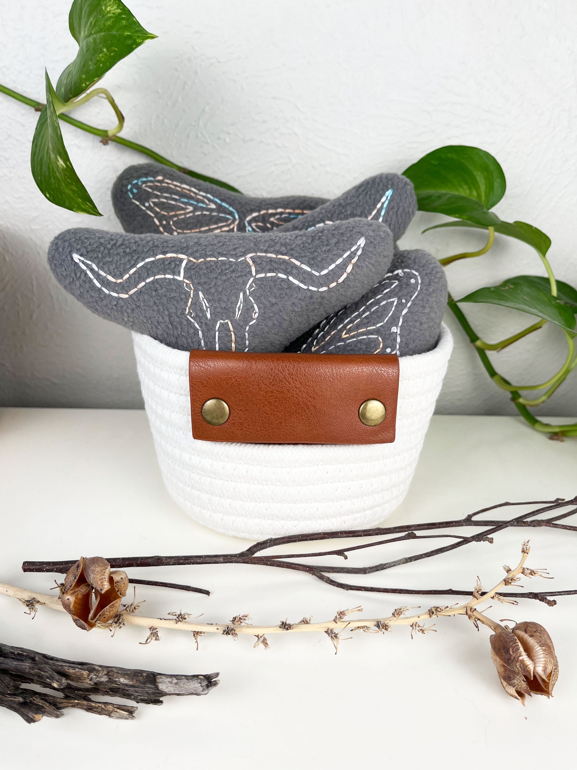 a group of grey stuffies hand embroidered in peach and sky blue variegated thread with longhorn skulls and butterfly wings in a white rope basket on a white counter with a pothos plant and dried twigs and yucca in front of it