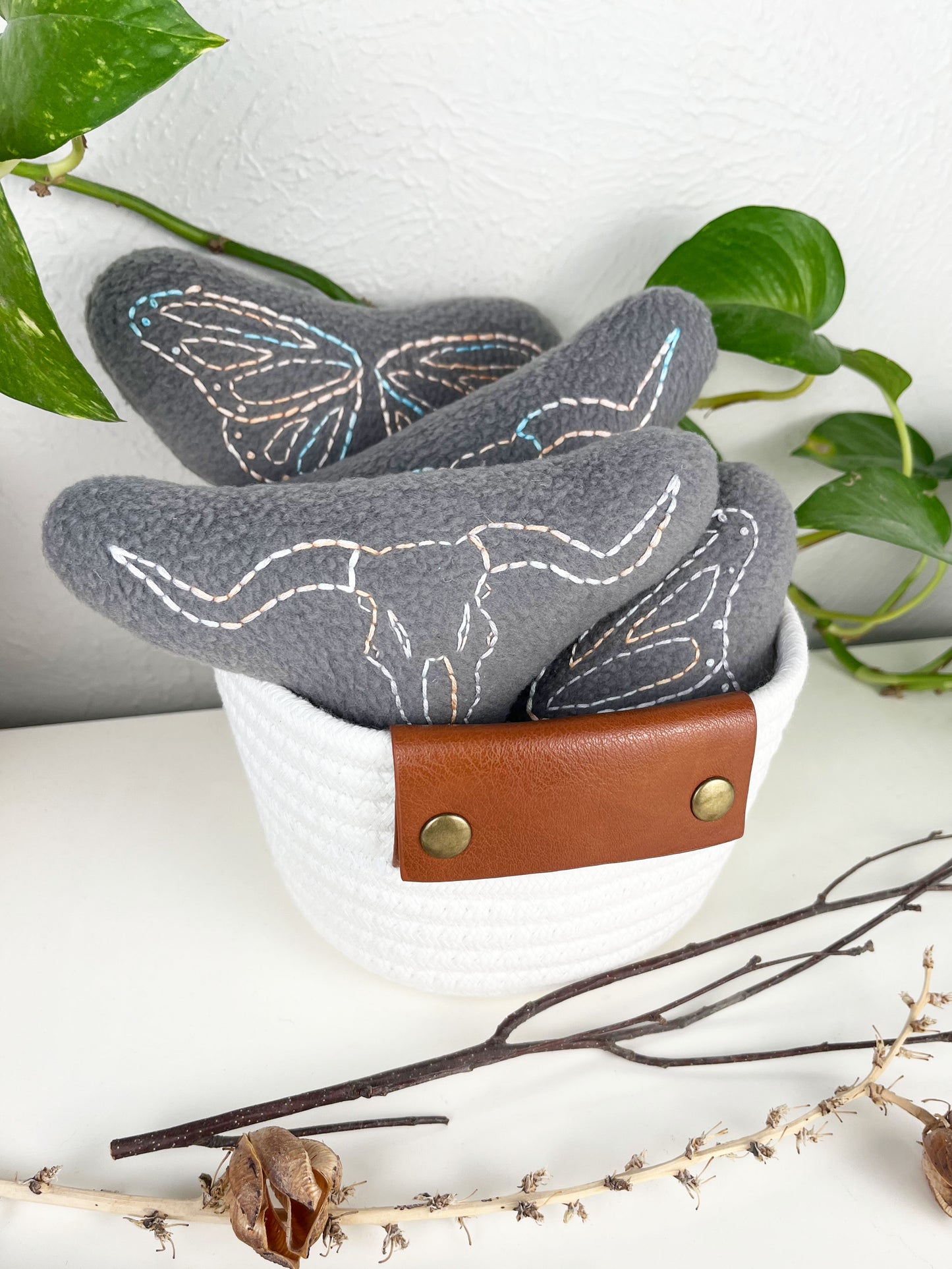 a group of grey stuffies hand embroidered in peach and sky blue variegated thread with longhorn skulls and butterfly wings in a white rope basket on a white counter with a pothos plant and dried twigs and yucca in front of it