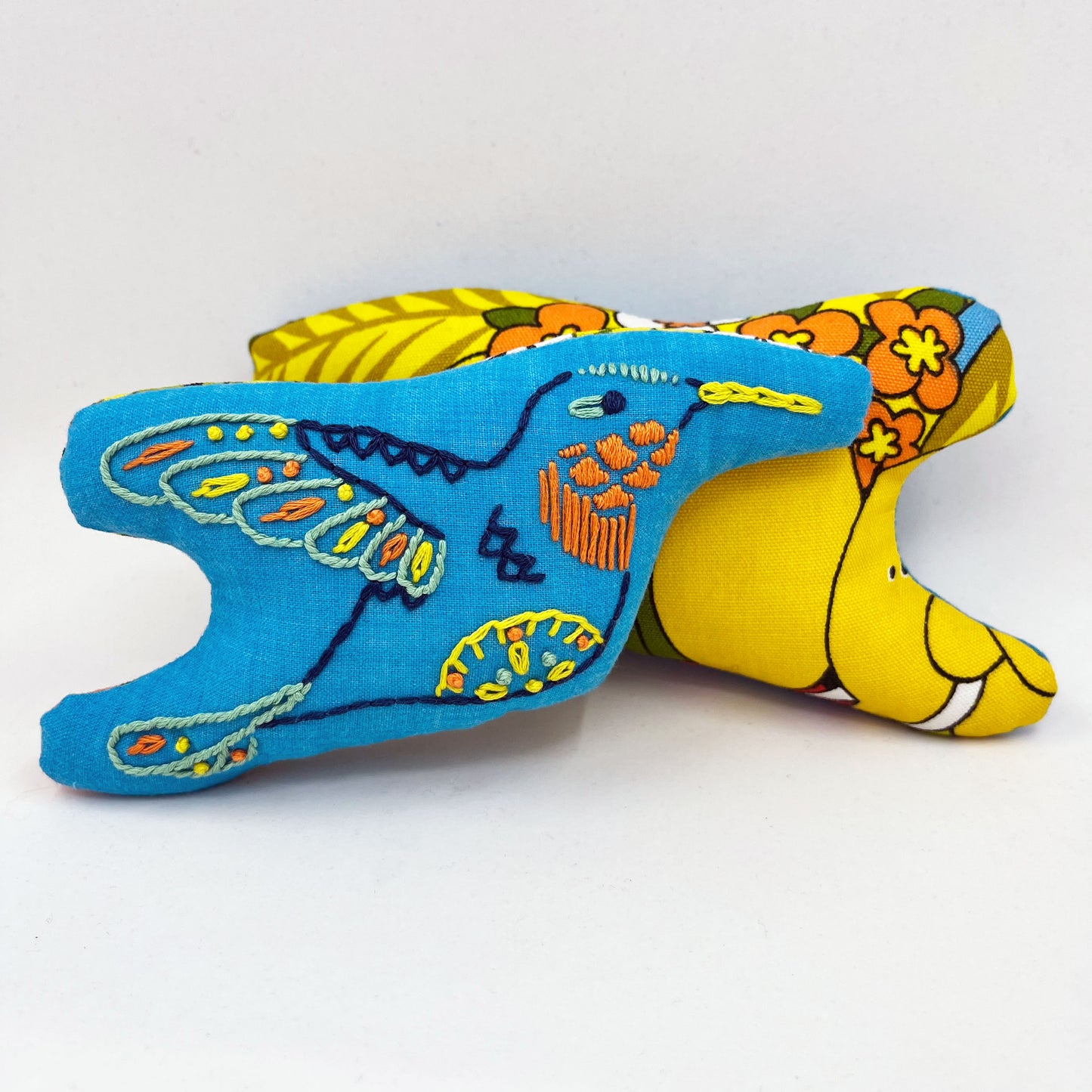 a colorfully hand embroidered stuffed pillow animal of a hummingbird on blue fabric, behind it one turned around showing the back side made of fabric with a cartoon jungle print