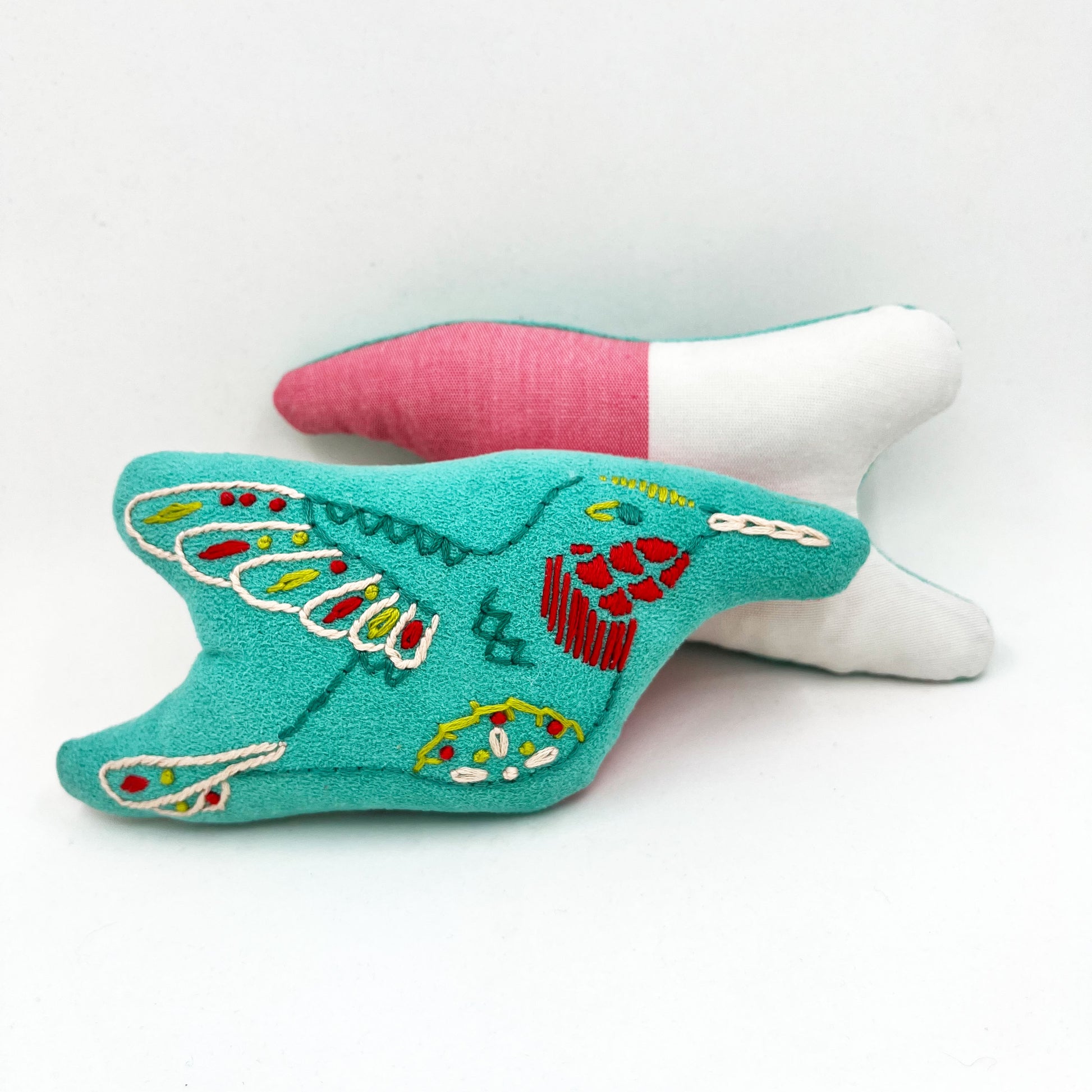 a colorfully hand embroidered stuffed pillow animal of a hummingbird on aqua fabric, behind it one turned around showing the back side made of half white half pink fabric