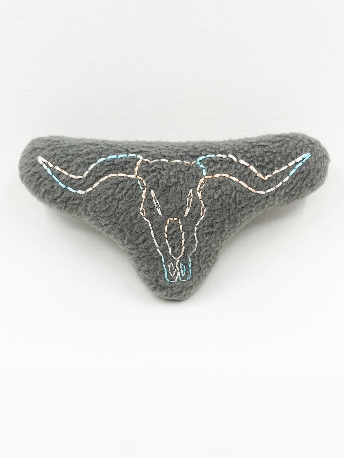  a grey fleece stuffed animal with a longhorn skull hand embroidered on it in peach and sky blue variegated thread with backstitch and french knots, on a white background