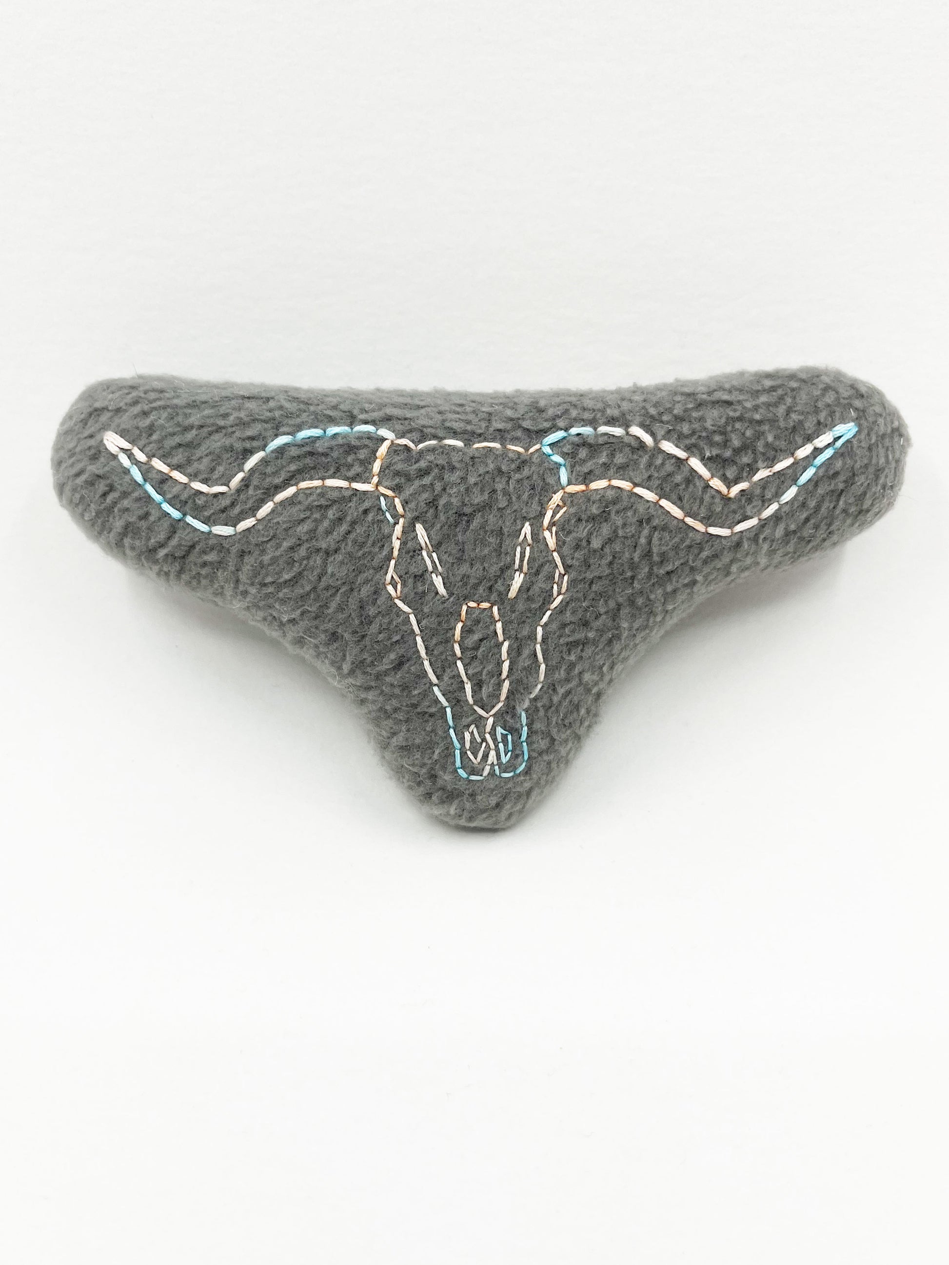  a grey fleece stuffed animal with a longhorn skull hand embroidered on it in peach and sky blue variegated thread with backstitch and french knots, on a white background