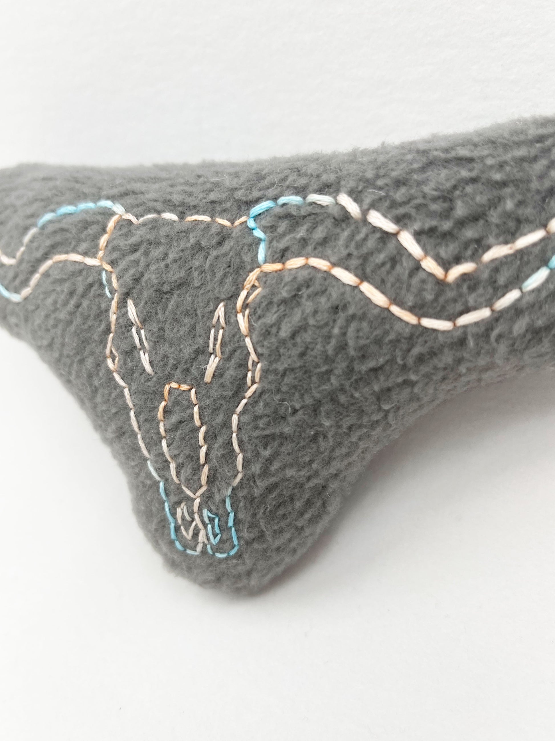 close up view of  a grey fleece stuffed animal with a longhorn skull hand embroidered on it in peach and sky blue variegated thread with backstitch and french knots, on a white background