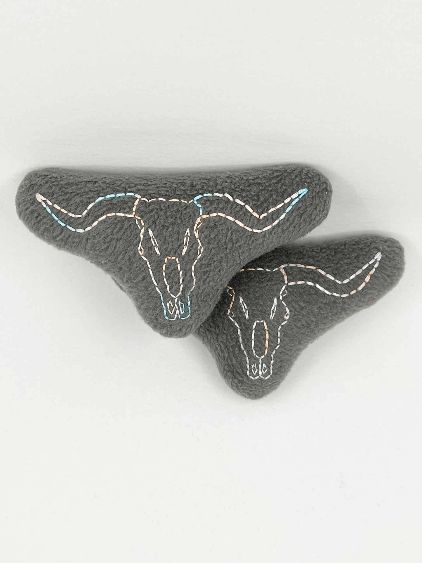 two grey fleece stuffed animals, with longhorn skulls hand embroidered on them in peach and sky blue variegated thread with backstitch and french knots, on a white background