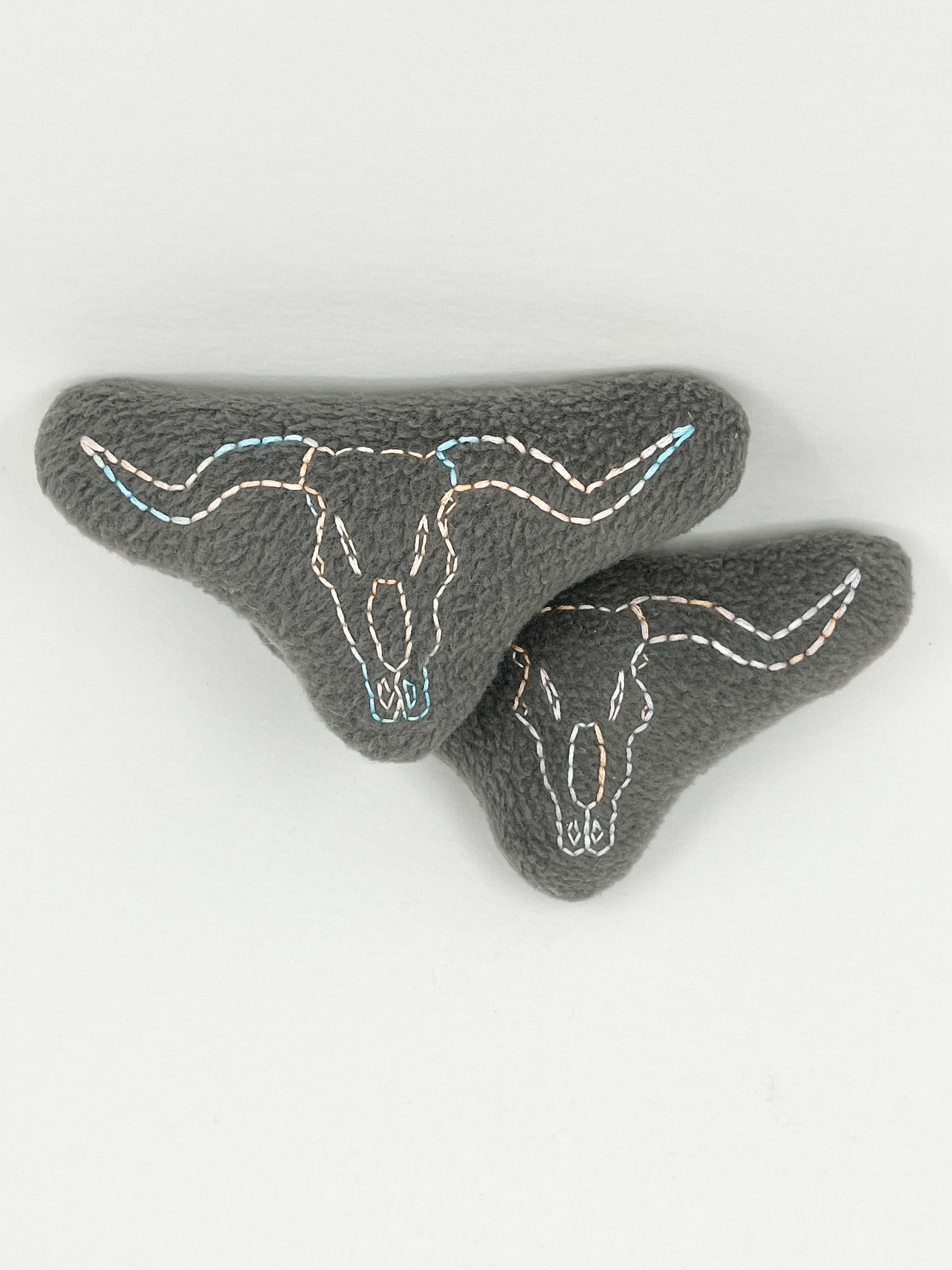 two grey fleece stuffed animals, with longhorn skulls hand embroidered on them in peach and sky blue variegated thread with backstitch and french knots, on a white background