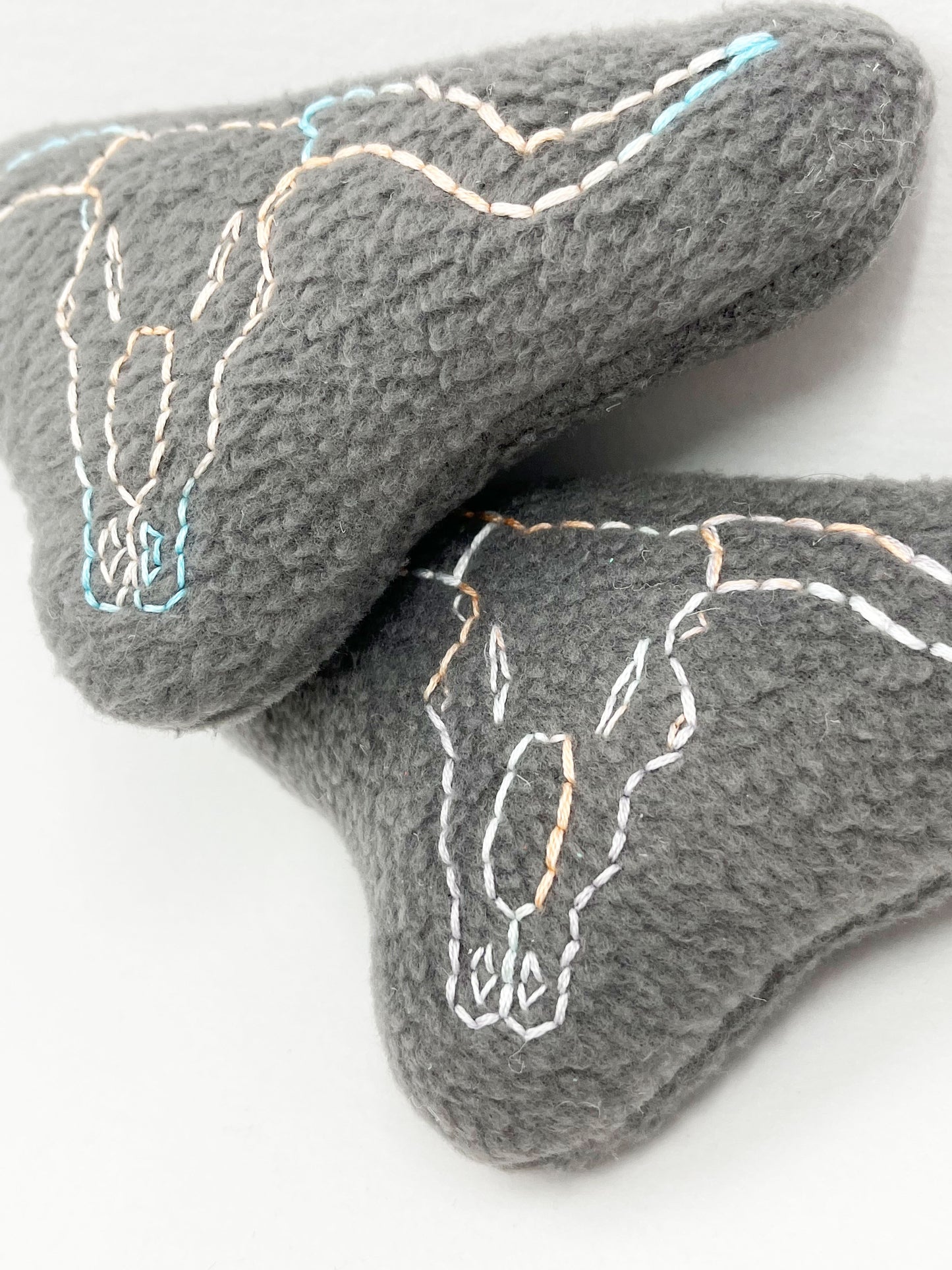 two grey fleece stuffed animals, with longhorn skulls hand embroidered on them in peach and sky blue variegated thread with backstitch and french knots, on a white background