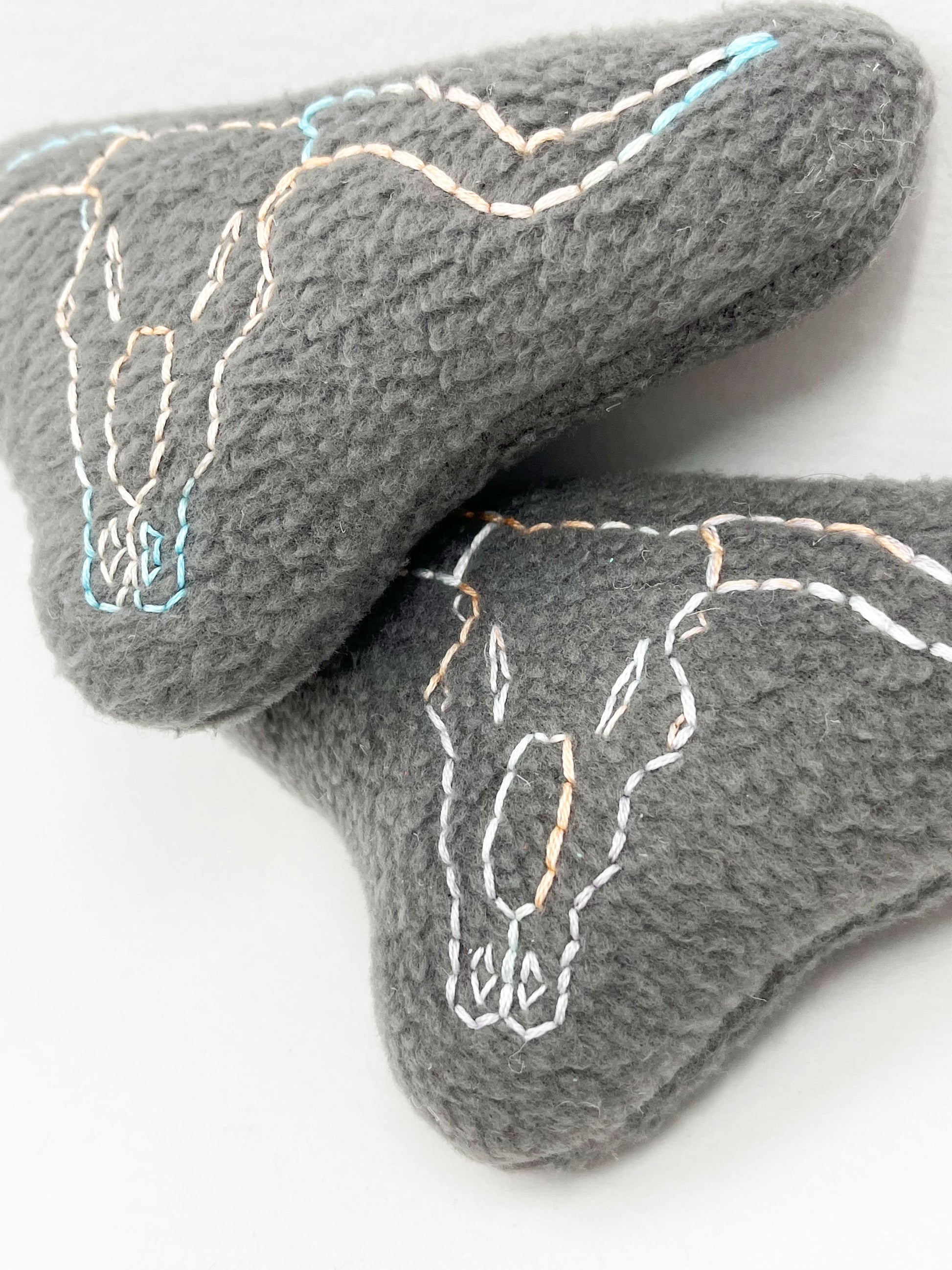 two grey fleece stuffed animals, with longhorn skulls hand embroidered on them in peach and sky blue variegated thread with backstitch and french knots, on a white background