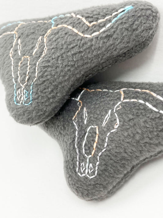 two grey fleece stuffed animals, with longhorn skulls hand embroidered on them in peach and sky blue variegated thread with backstitch and french knots, on a white background