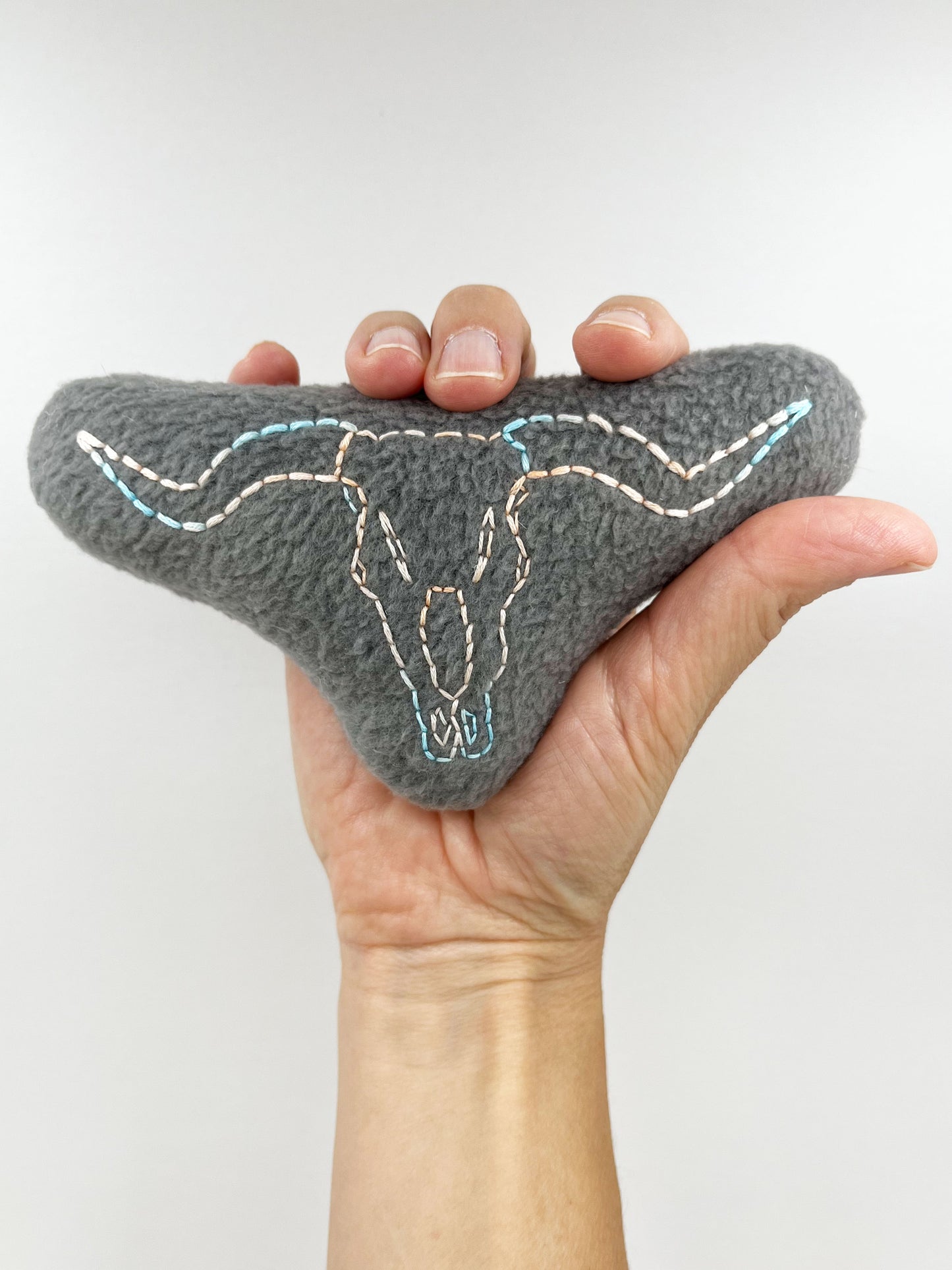 a hand holding up a grey fleece stuffed animal with a longhorn skull hand embroidered on it in peach and sky blue variegated thread with backstitch and french knots