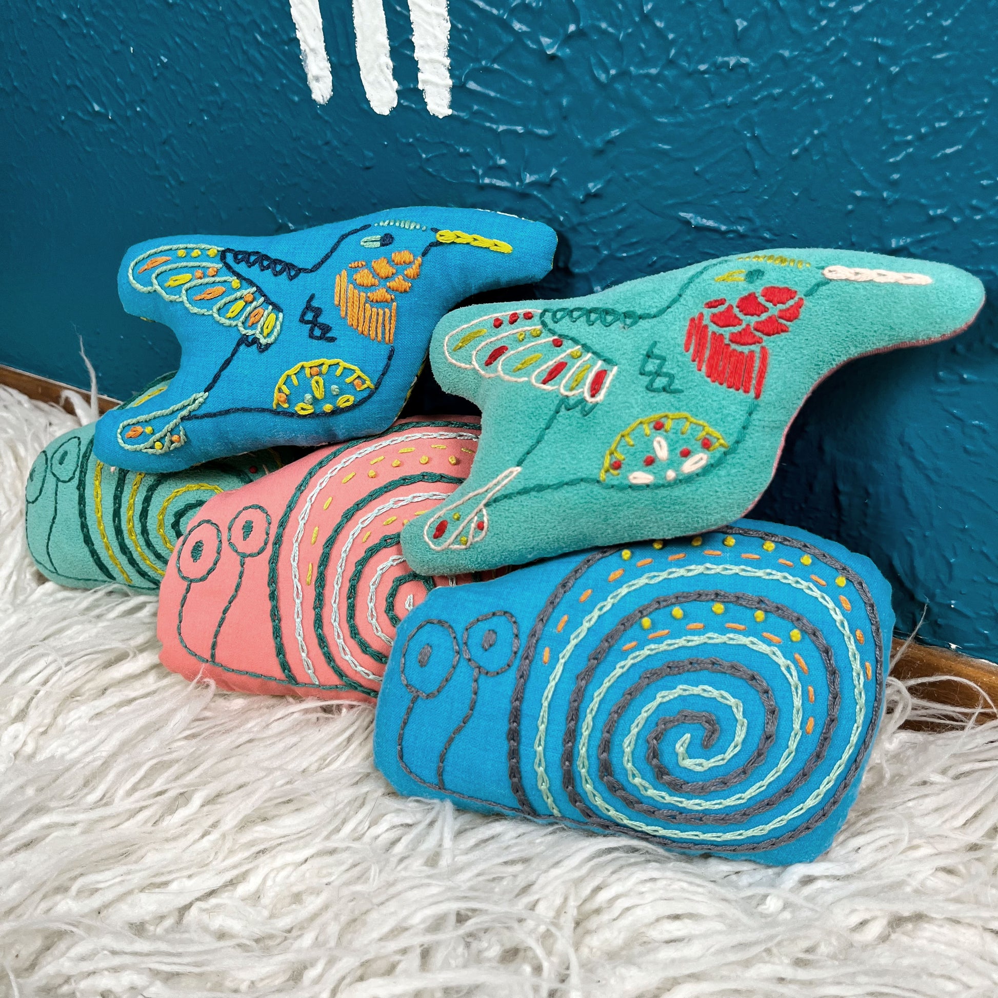 a group of colorfully hand embroidered hummingbird and snail stuffed animal pillows in blue, seafoam green and coral fabric, in a pile on a furry white rug in front of a teal wall
