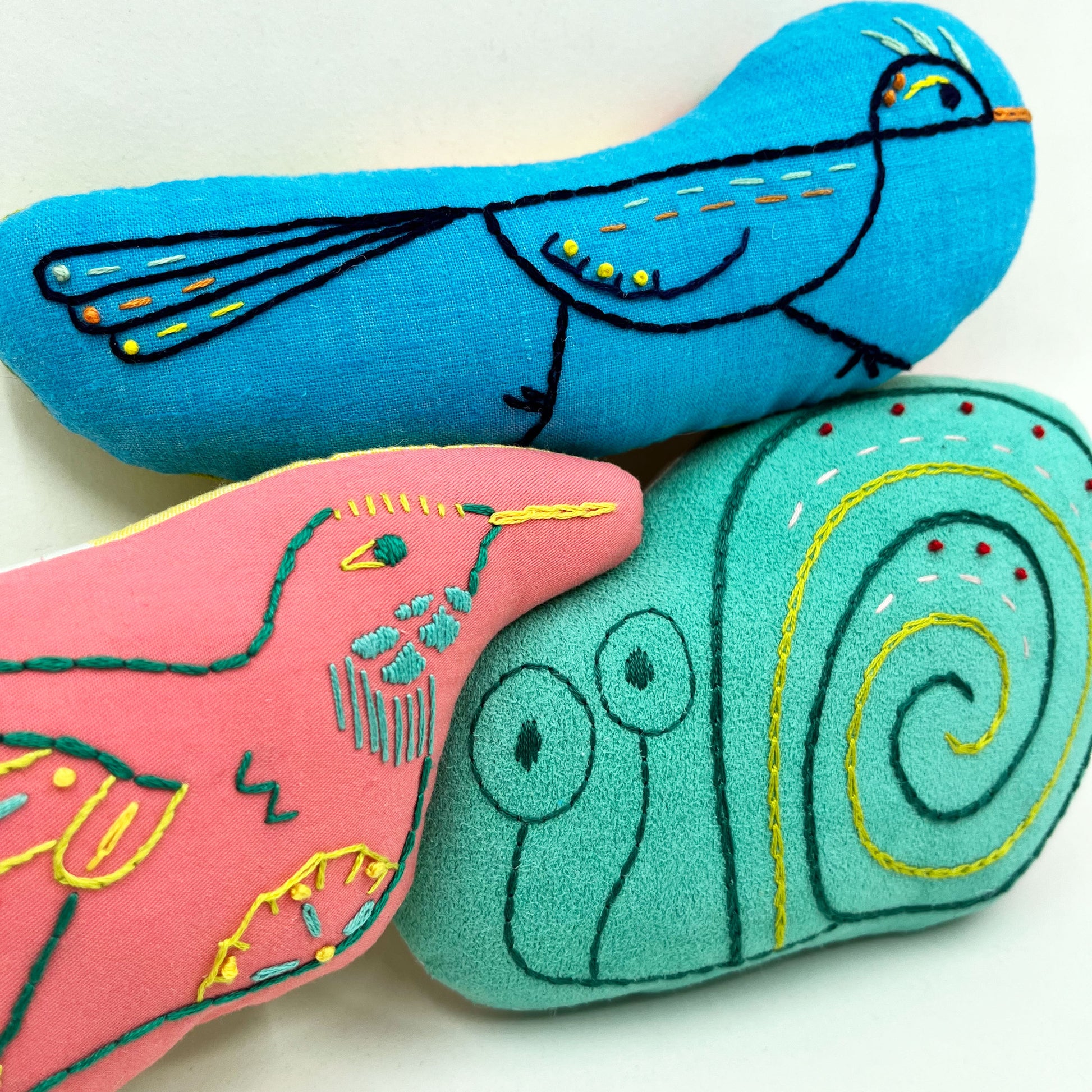 a group of colorfully hand embroidered stuffed pillow animals-a roadrunner, snail and hummingbird in coral, aqua and blue fabric,  with a white background