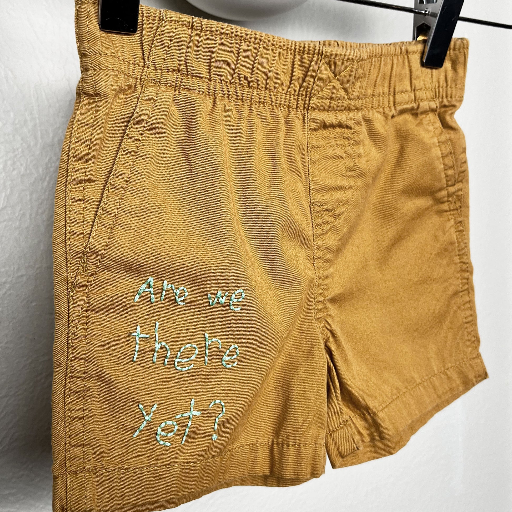 close up view of small camel colored shorts hanging from a wooden hanger, the words "are we there yet?" are hand embroidered on the lower right leg