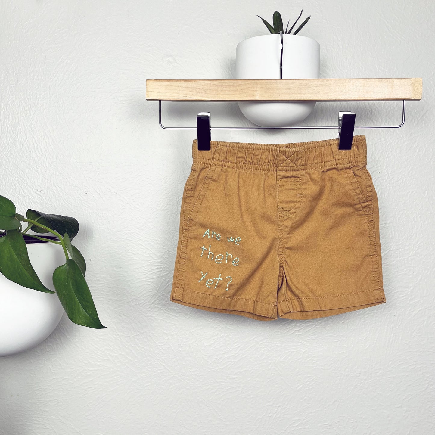 small camel colored shorts hanging from a wooden hanger, the words "are we there yet?" are hand embroidered on the lower right leg, there is a pothos plant in a round white pot hanging on the wall near the shorts