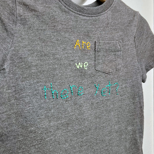 Close up view of a grey short sleeve pocket tee, with the words "Are we there yet?" hand embroidered on it in mustard and shades of green