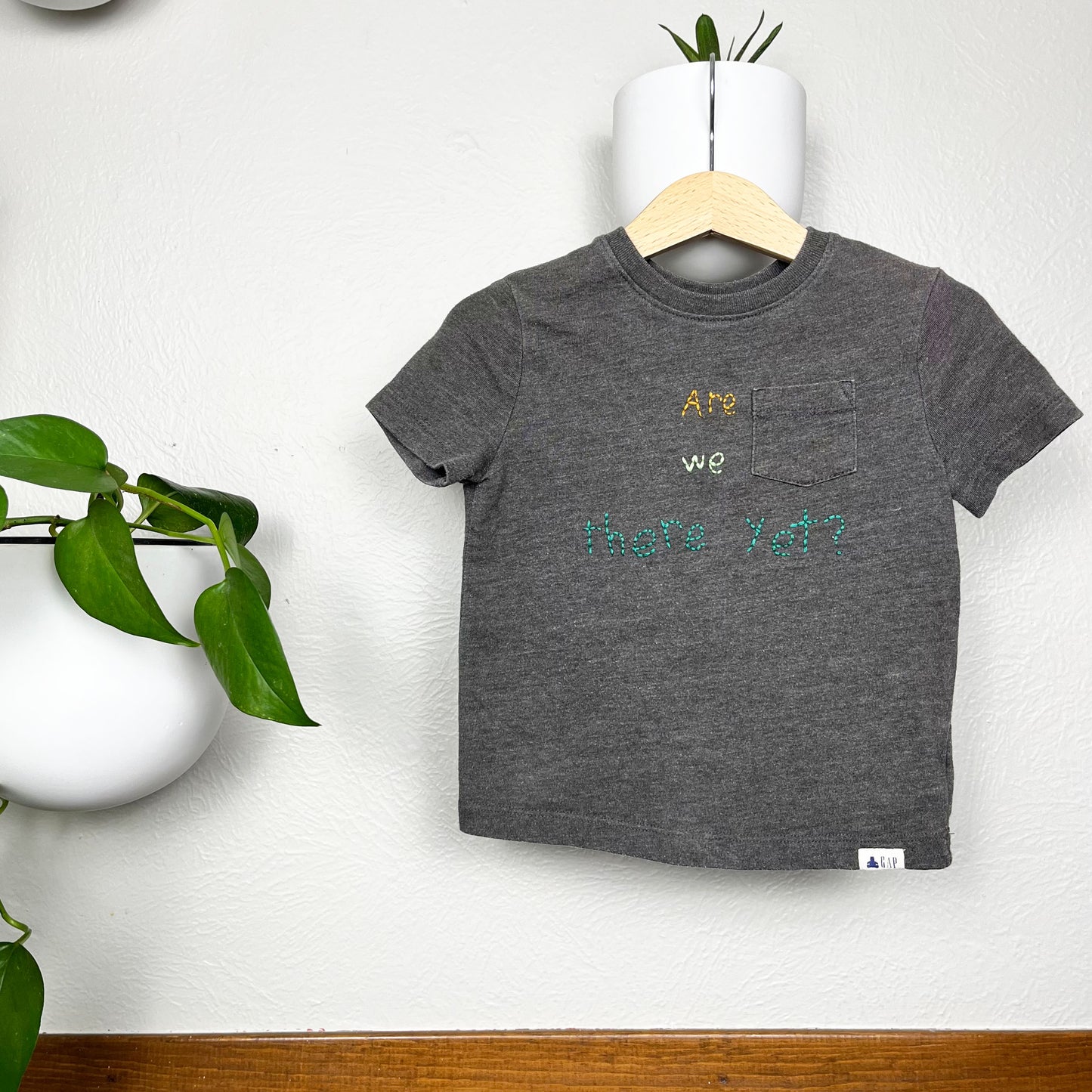 a grey short sleeve pocket tee, with the words "Are we there yet?" hand embroidered on it in mustard and shades of green, on a hanger, hanging off a white round planter on the wall with another plant on the wall next to it