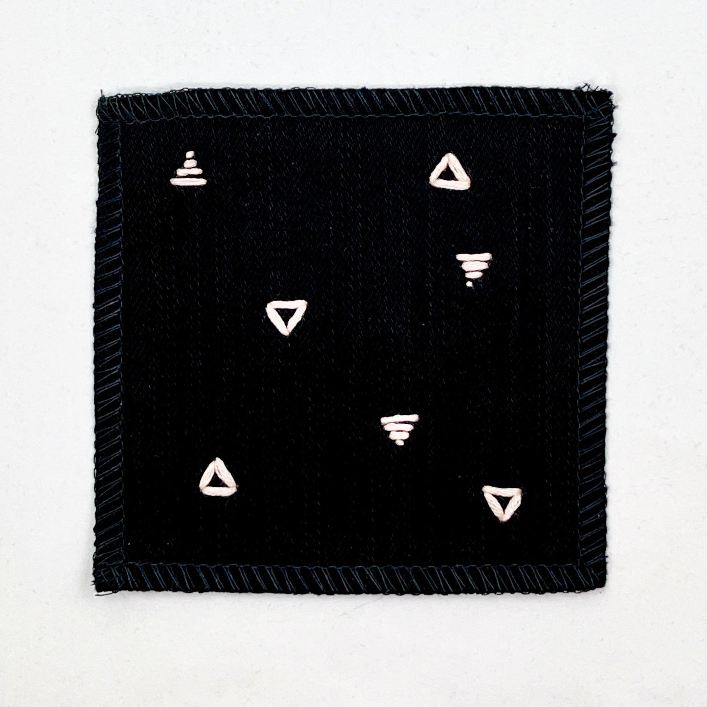 a square patch made out of black canvas, handstitched with scattered peach triangles, some as outlines some with a line fill, with overlocked edges, on a white background