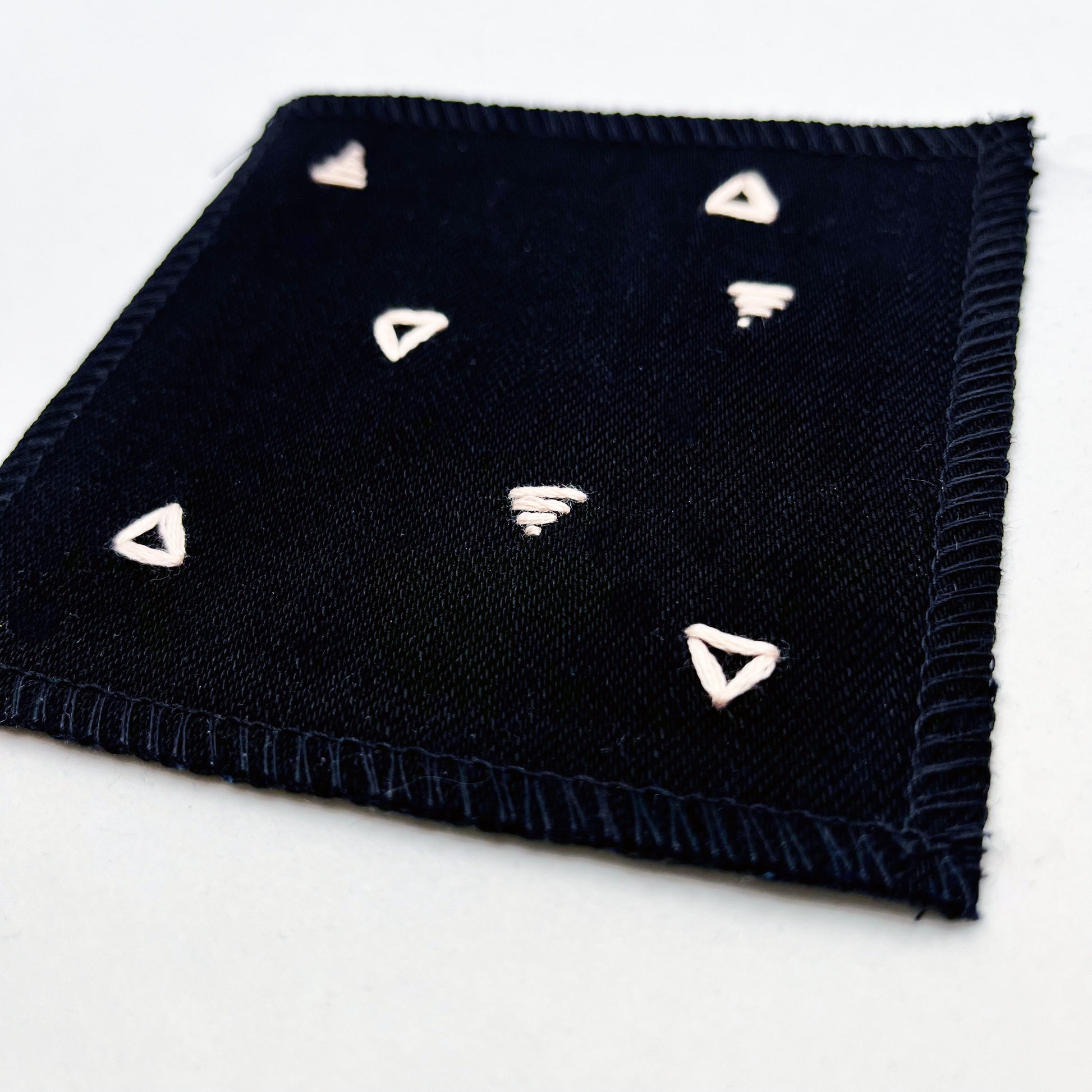 close up angled view of a square patch made out of black canvas, handstitched with scattered peach triangles, some as outlines some with a line fill, with overlocked edges, on a white background