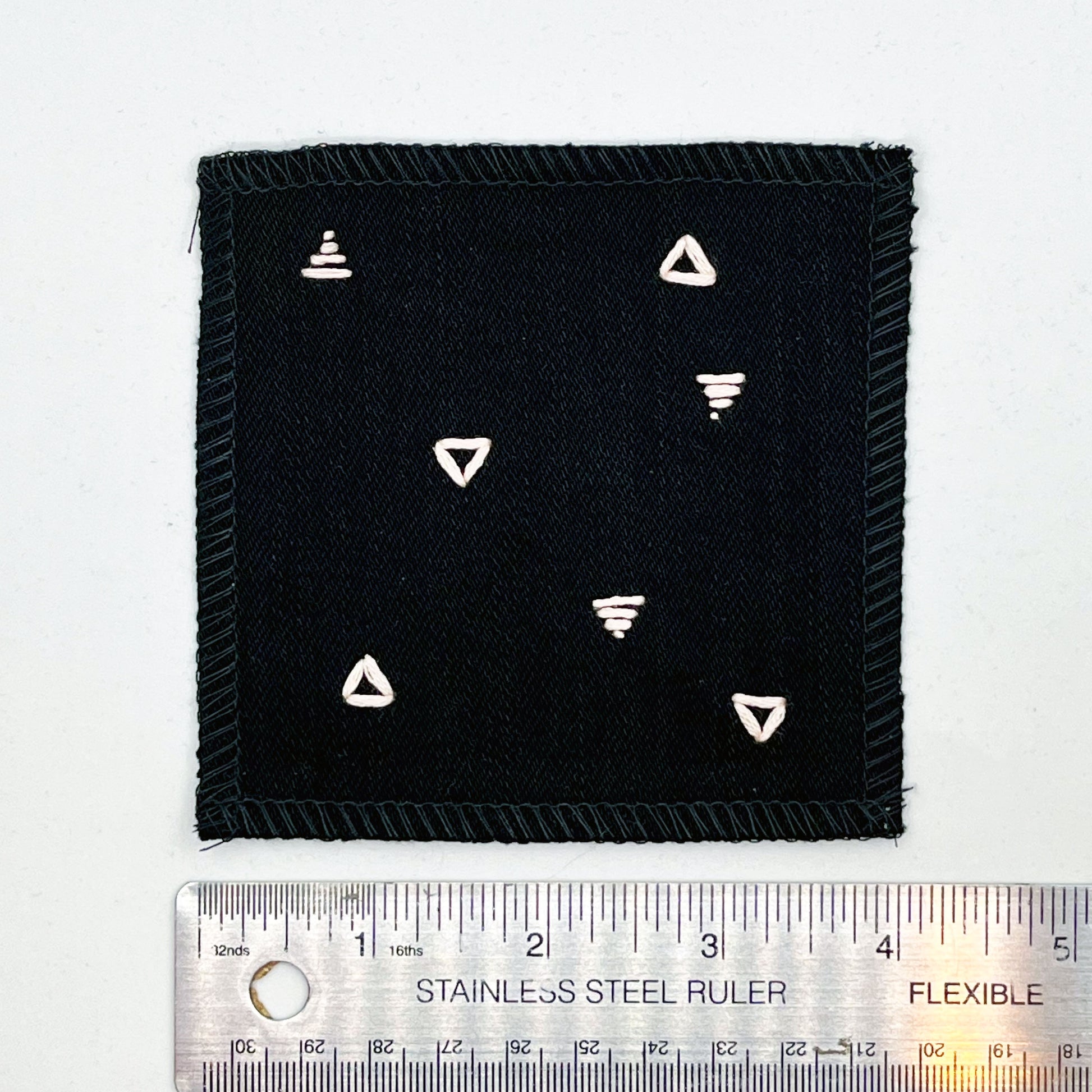 a square patch made out of black canvas, handstitched with scattered peach triangles, some as outlines some with a line fill, with overlocked edges, on a white background, next to a ruler showing a width of 4 inches