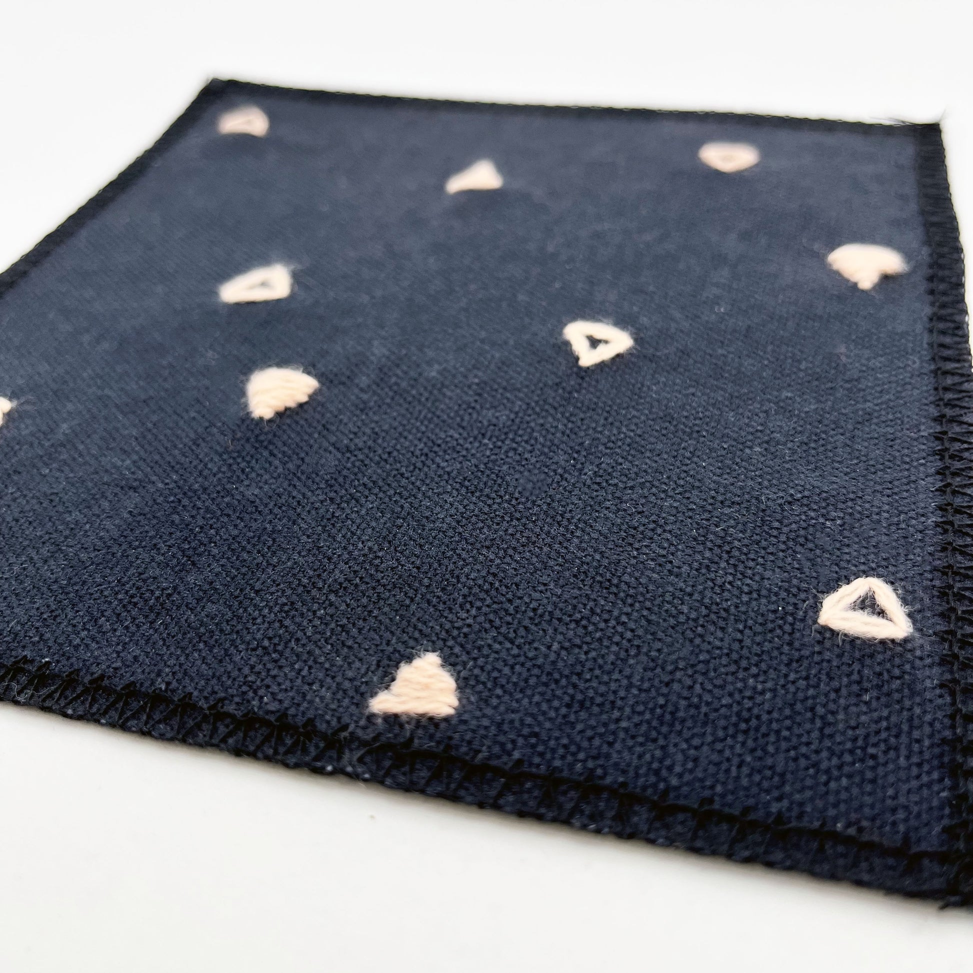 close up angled view of a square patch made out of black canvas, handstitched with scattered peach triangles, some as outlines some with a line fill, with overlocked edges, on a white background