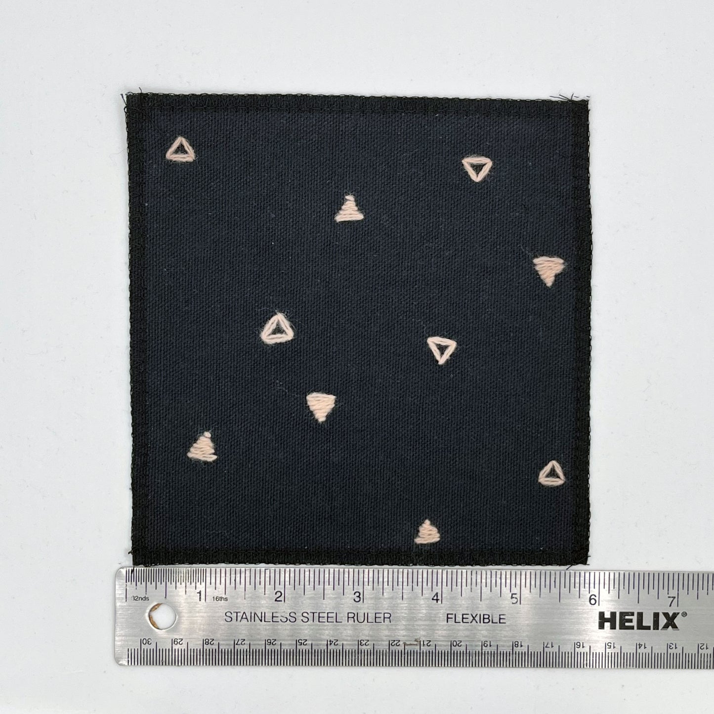 a square patch made out of black canvas, handstitched with scattered peach triangles, some as outlines some with a line fill, with overlocked edges, on a white background, next to a ruler showing a width of 6 inches